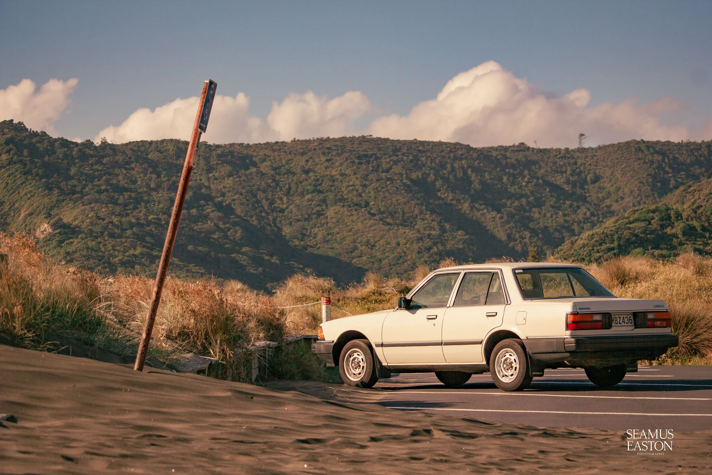 A vintage white sedan parked near a sandy area with grass, with lush green hills and a partly cloudy sky in the background.