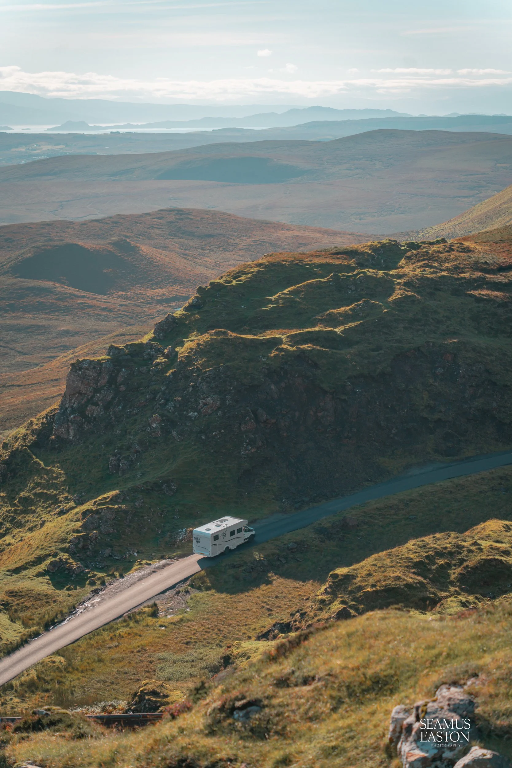 A white camper van driving on a narrow paved road through green hilly landscape with distant mountains and partly cloudy sky.