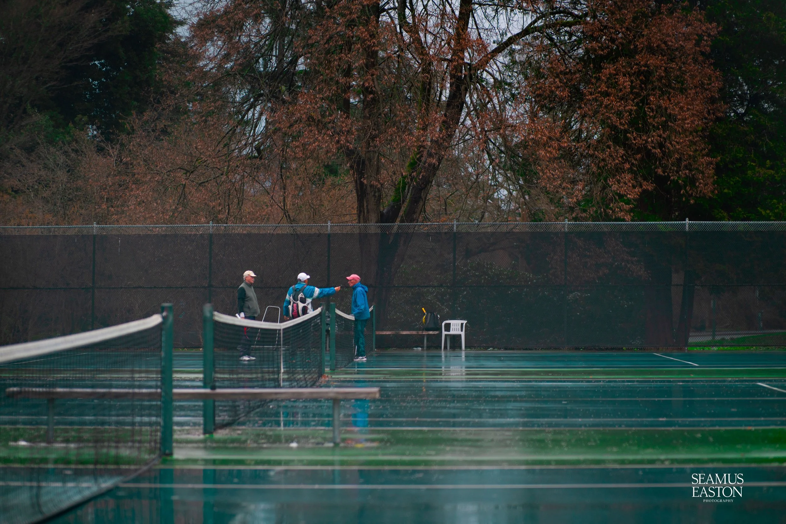 Four people standing on a wet tennis court near a black fence, chatting, with leafless trees in the background and an overcast sky.