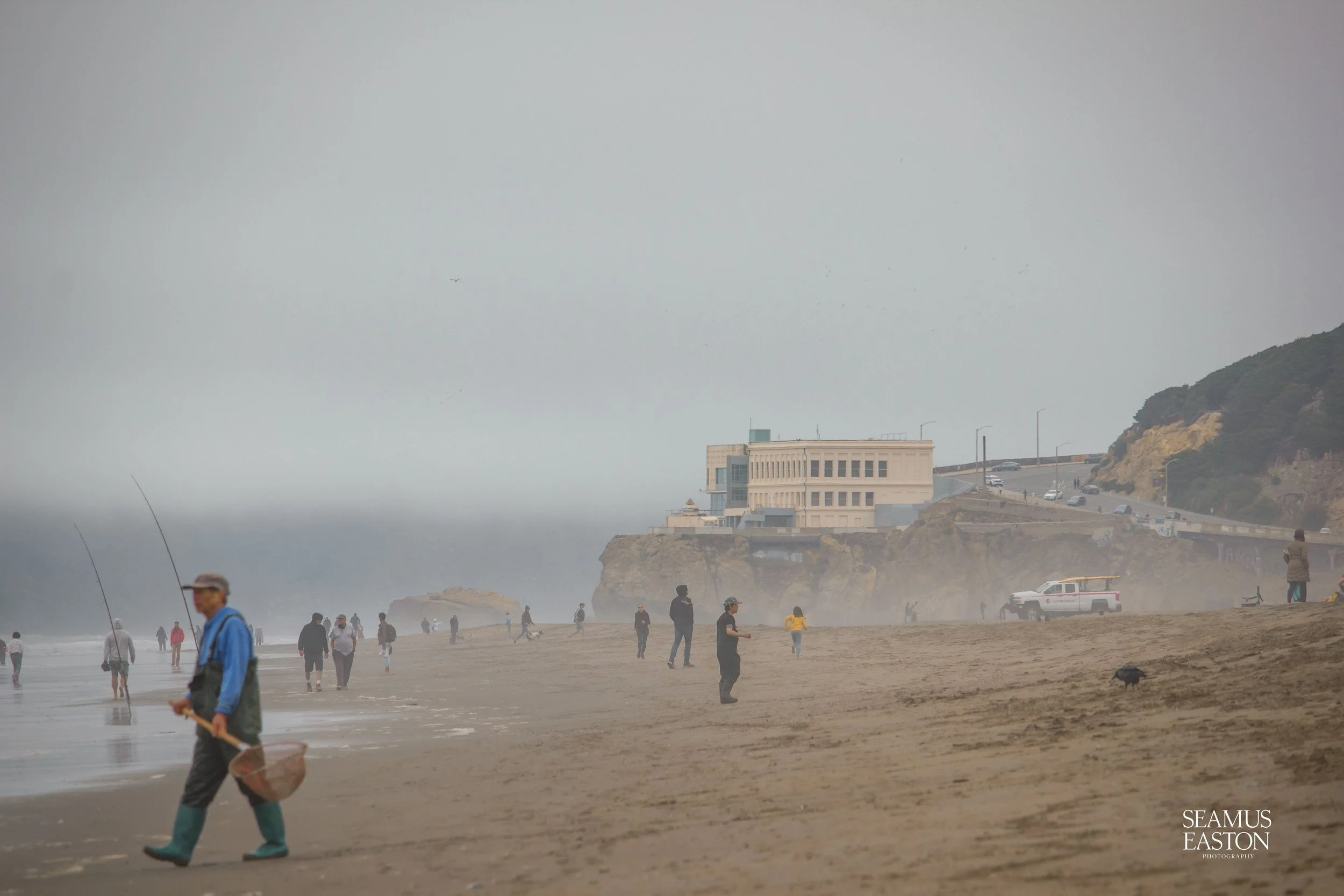 People fishing on a foggy beach with a large building on a cliff in the background and a misty sky overhead.