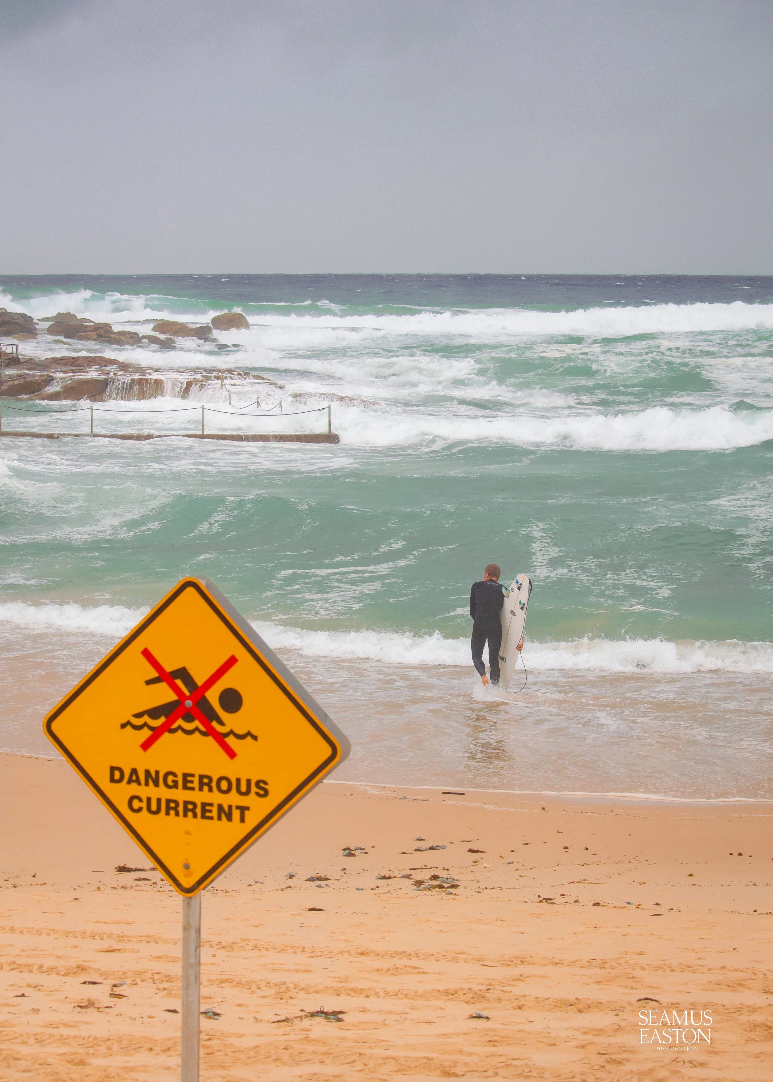 A person with a surfboard walking into the ocean at a beach with a yellow warning sign that reads 'Dangerous Current' and depicts a swimmer crossed out.