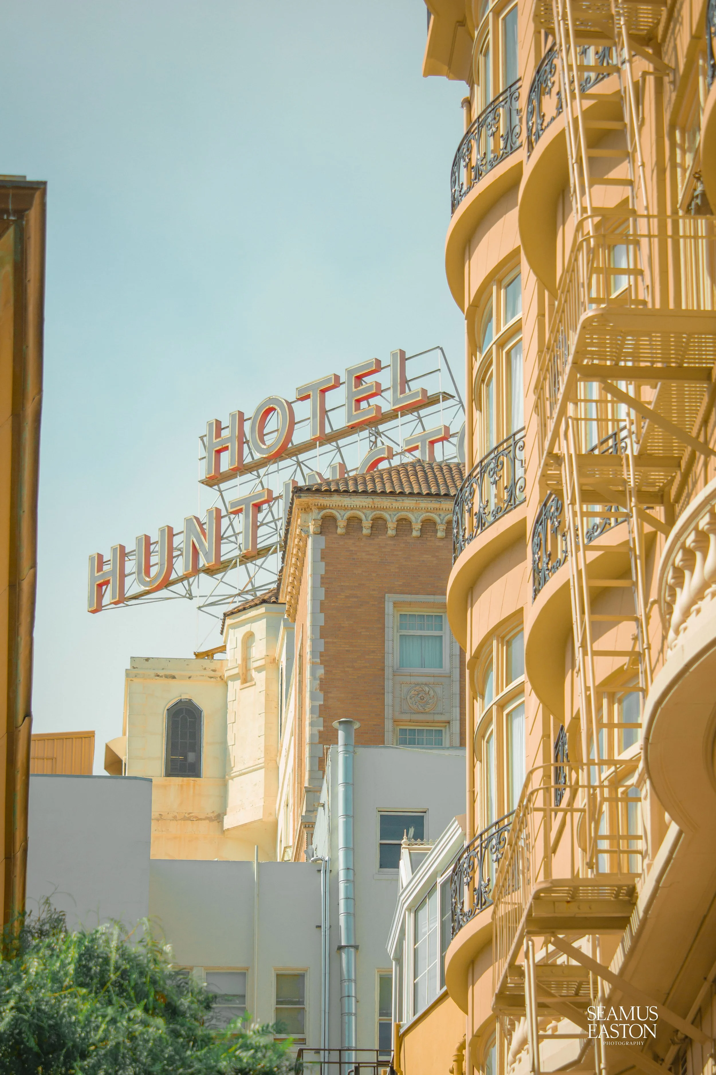 An upward view of a hotel building with orange and white exterior, decorative balconies, and fire escapes. A large, vintage hotel sign reads"HOTEL HUNTINGTON" against a clear blue sky.