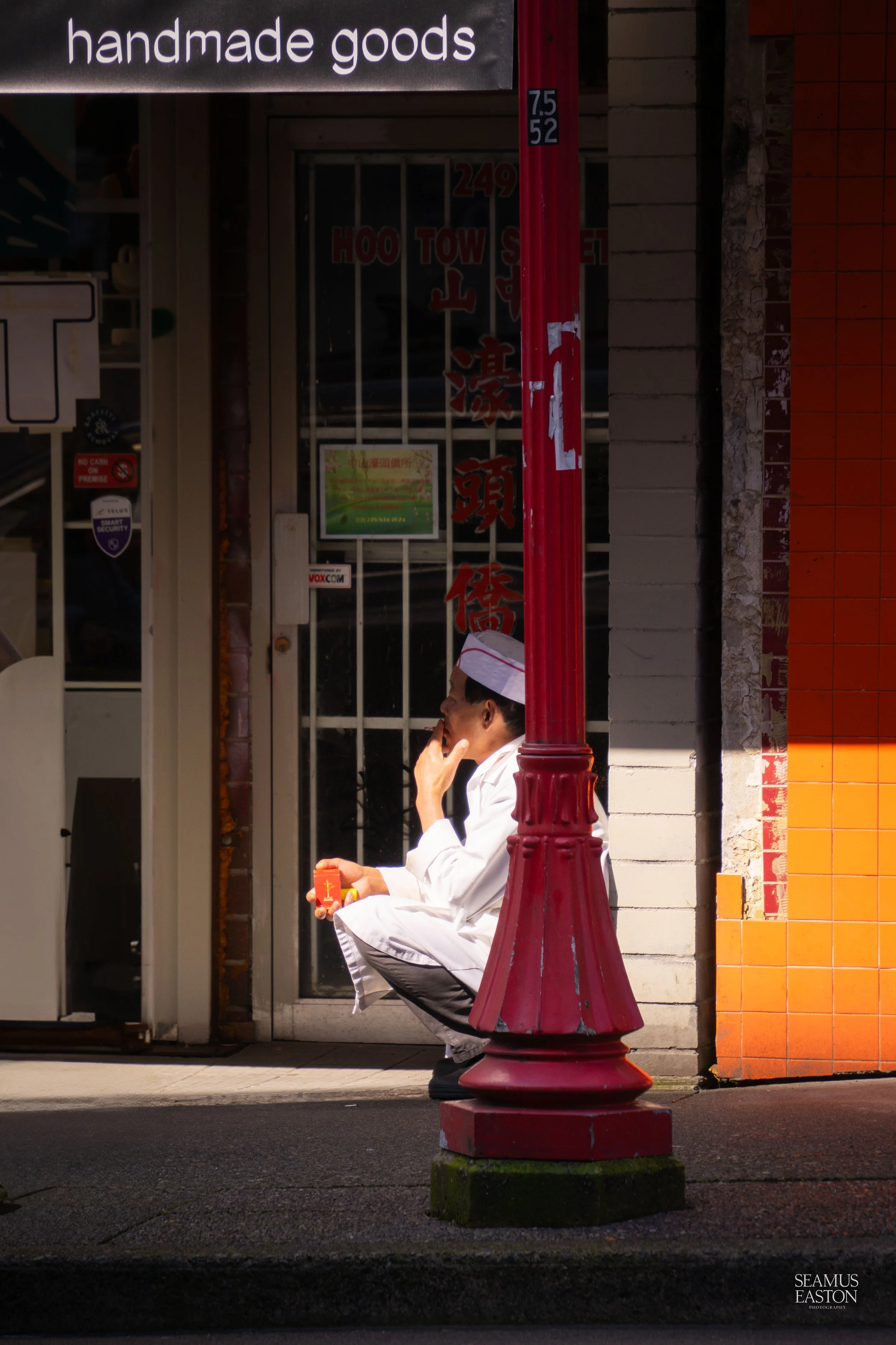 A man dressed as a chef sitting on a sidewalk bench, smoking a cigarette and holding a small object, with a red lamppost in front of him. The scene is outside a store with a sign that reads 'handmade goods' and some Chinese characters on the window.