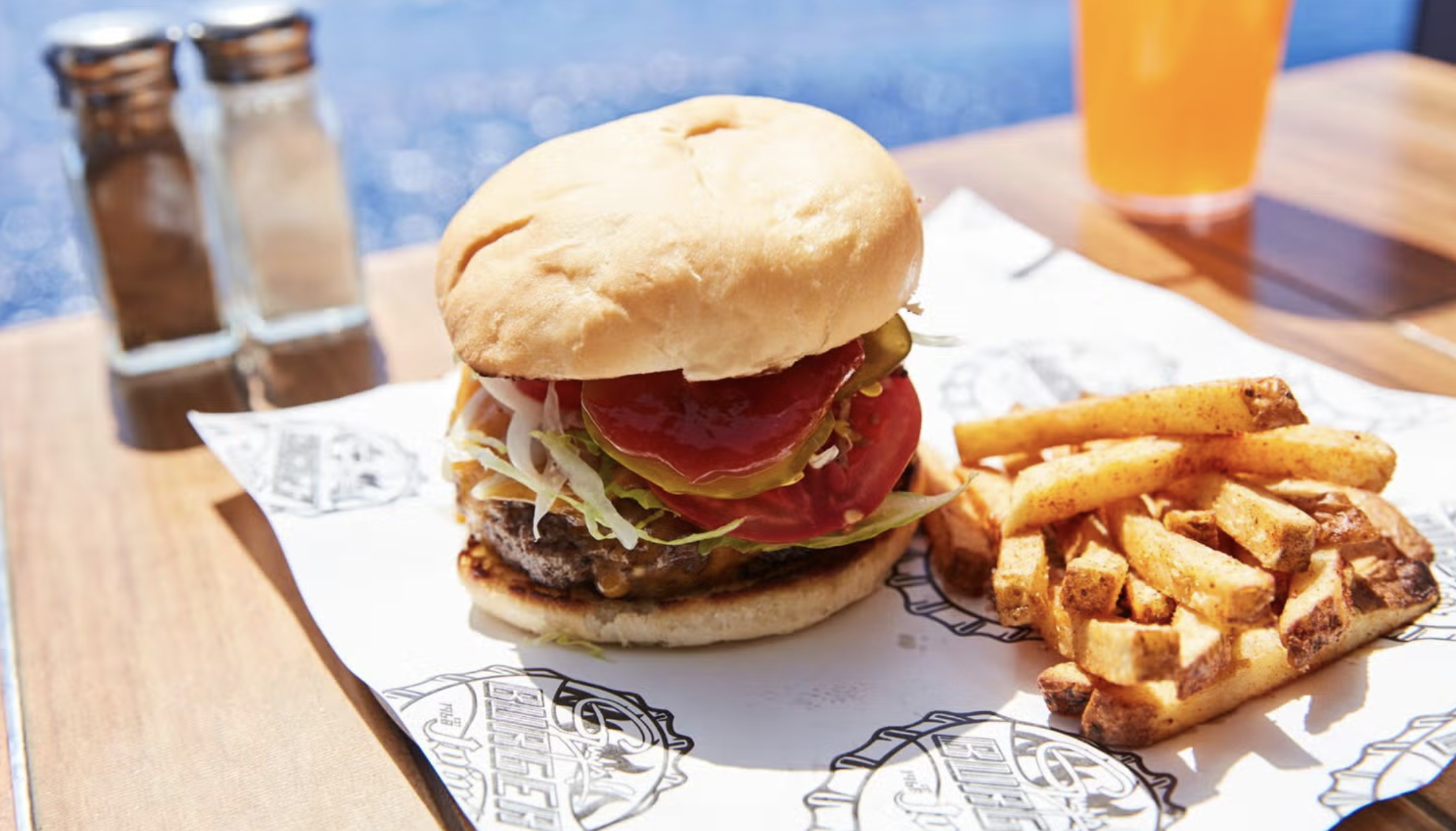 Burger topped with lettuce, tomato, pickles, and sauce beside seasoned fries on Guy’s Burger Joint paper.