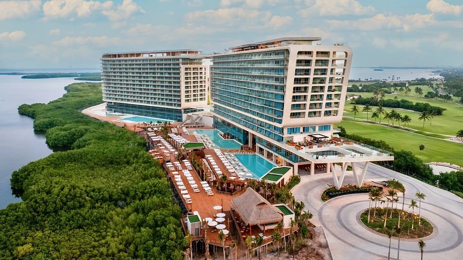Aerial view of a modern luxury resort with multiple pools, waterfront mangroves, and a neighboring golf course in Cancun.
