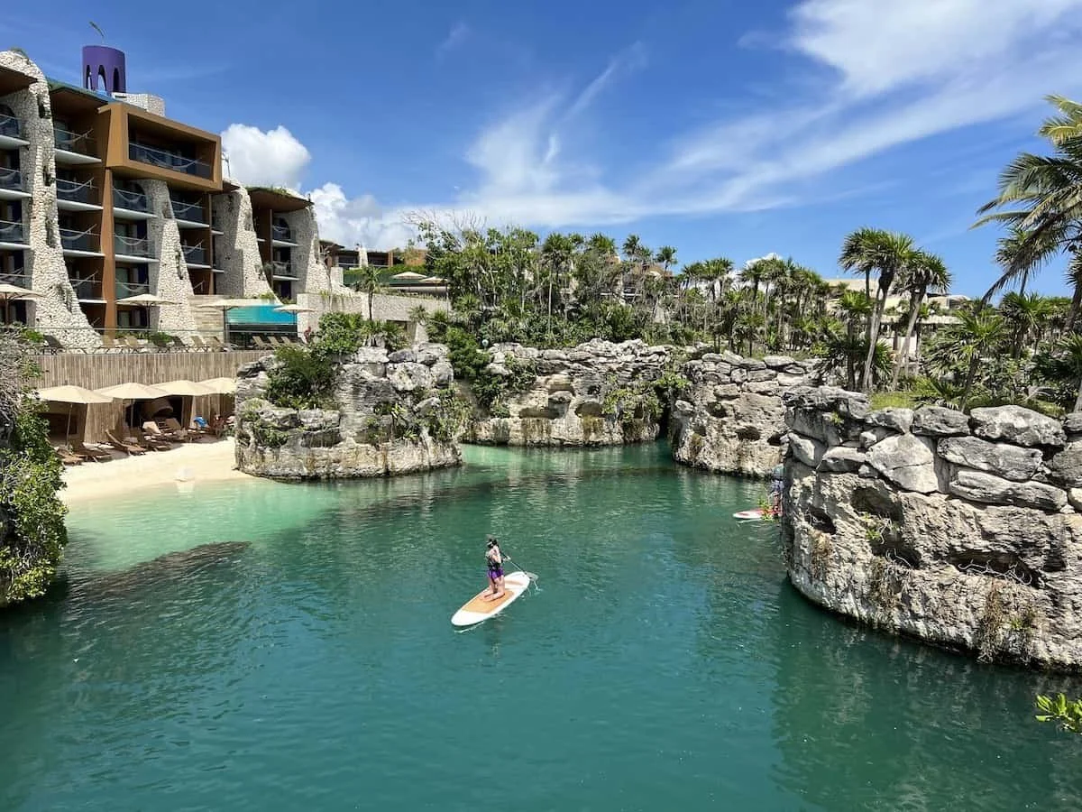 Guest paddleboarding through a turquoise lagoon surrounded by tropical greenery and limestone architecture at Hotel Xcaret Arte.