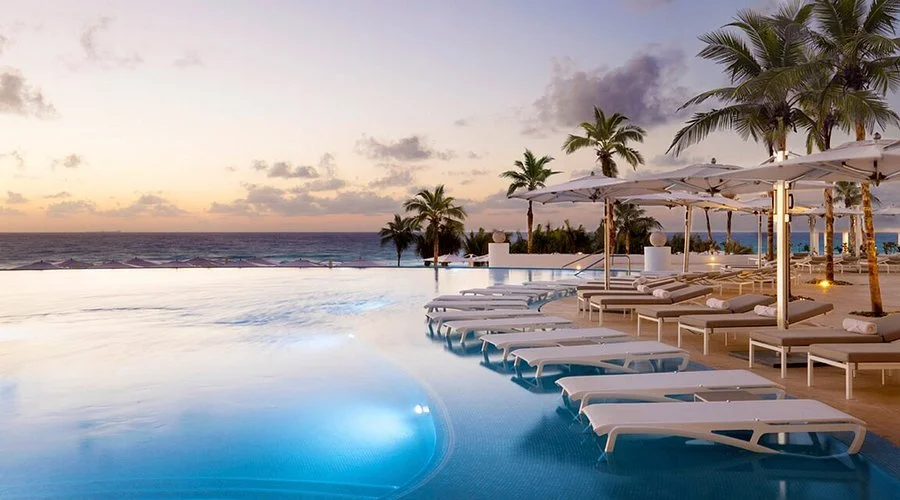Oceanfront infinity pool surrounded by lounge chairs, umbrellas, and palm trees at sunset at a luxury adults-only resort.