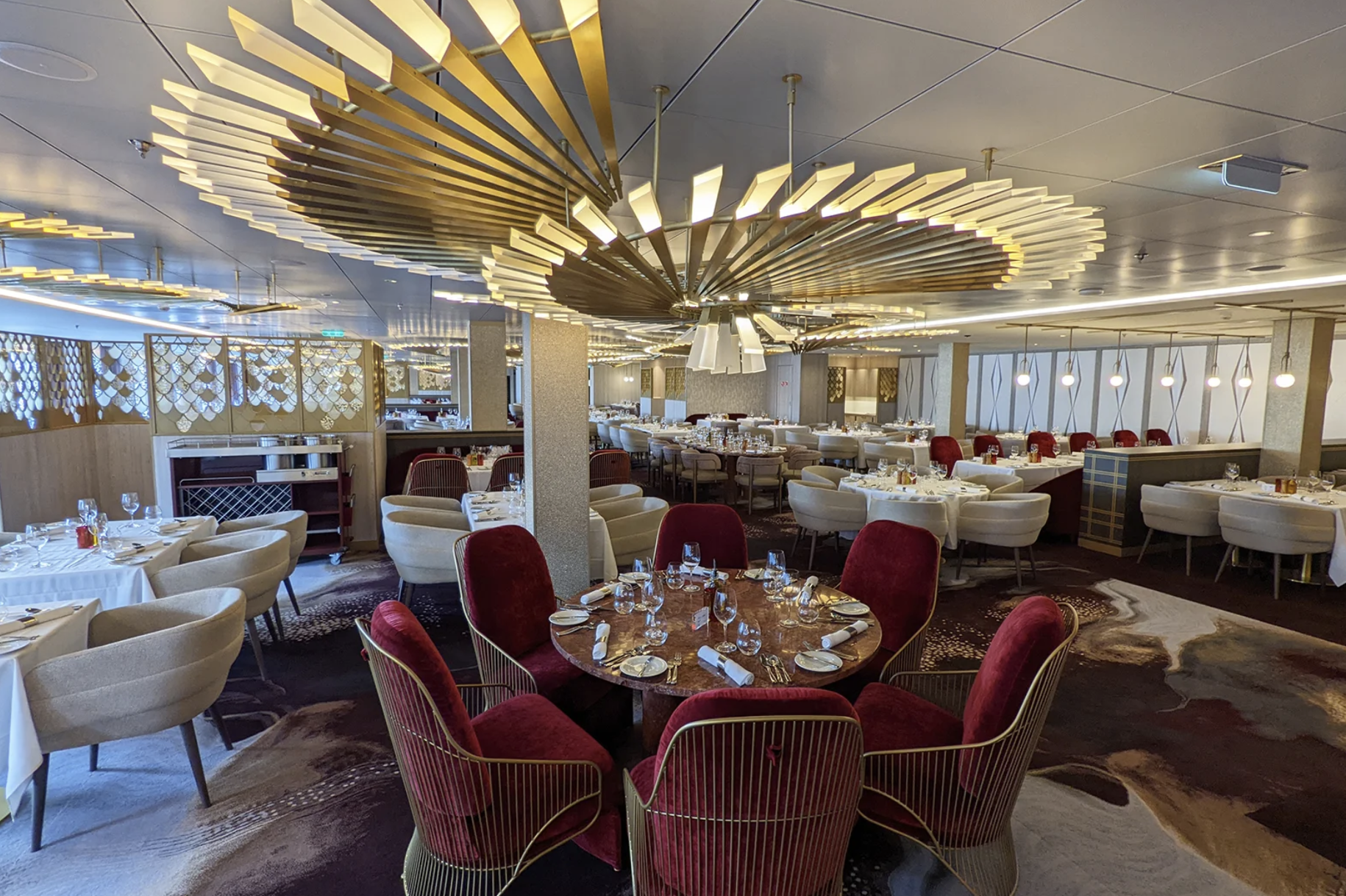 Celebrity Beyond dining room with red and cream chairs, white tablecloths, and sculptural gold chandeliers.