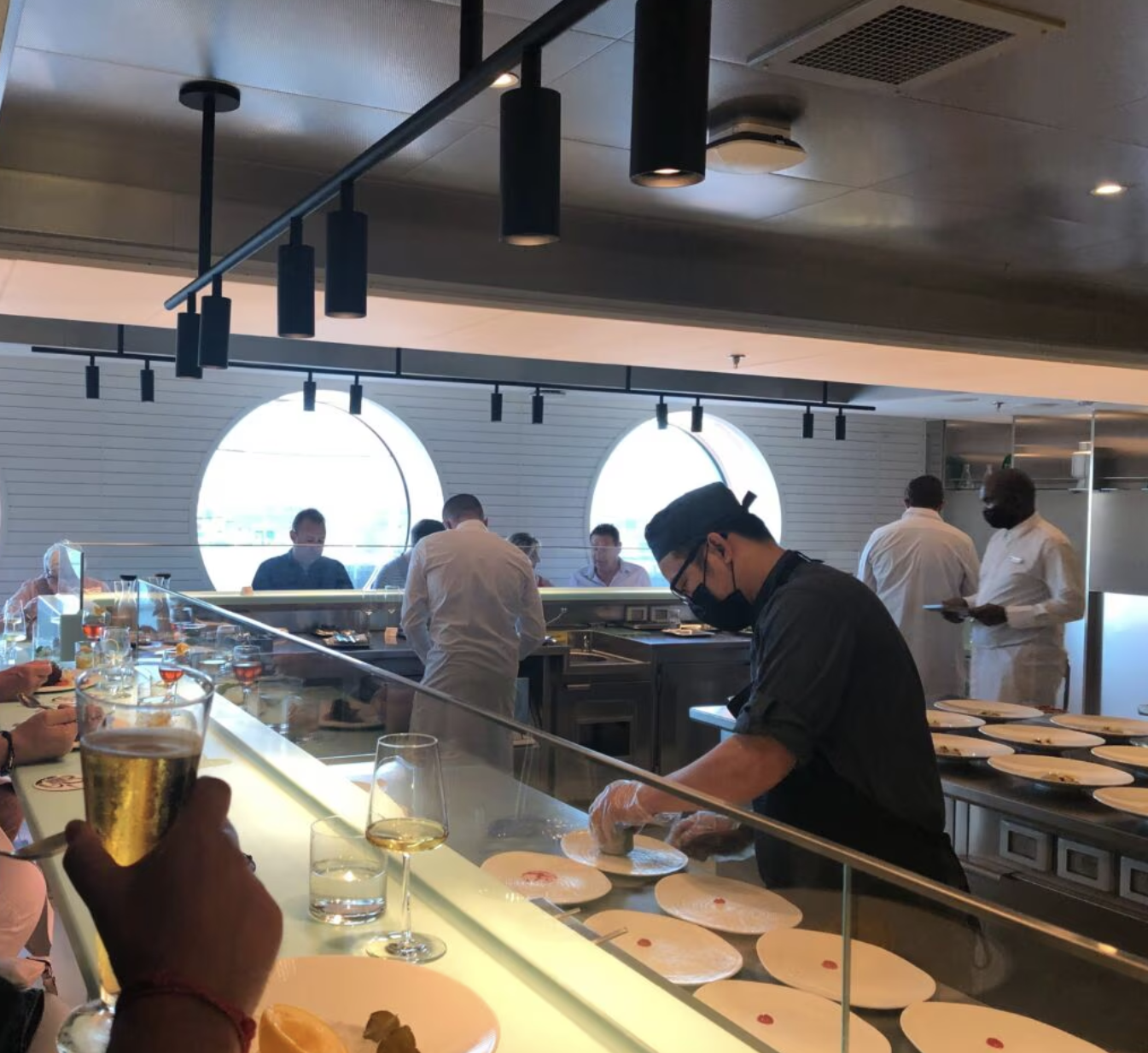 Chef prepares plated dishes behind a glass counter while guests watch in Scarlet Lady’s Test Kitchen.
