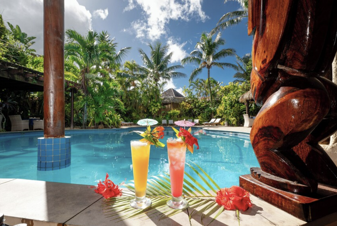 Two colorful tropical cocktails with umbrellas and flowers sitting beside a resort pool surrounded by palm trees