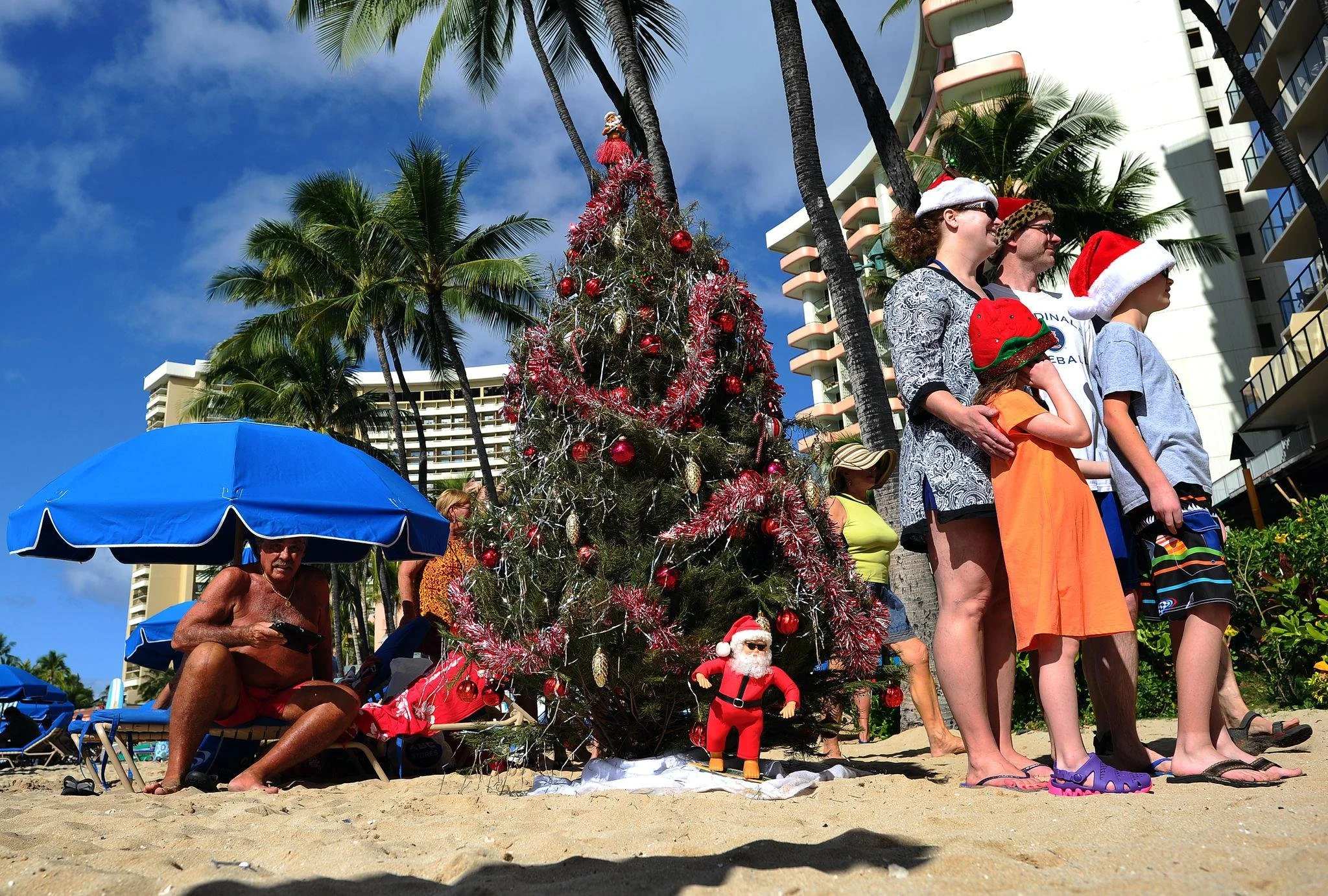 Family celebrating Christmas on a Hawaiian beach beside a decorated tree and palm trees.