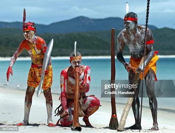 Three Ngaro performers in traditional body paint play the didgeridoo and hold ceremonial instruments on a beach in the Whitsundays, Australia.
