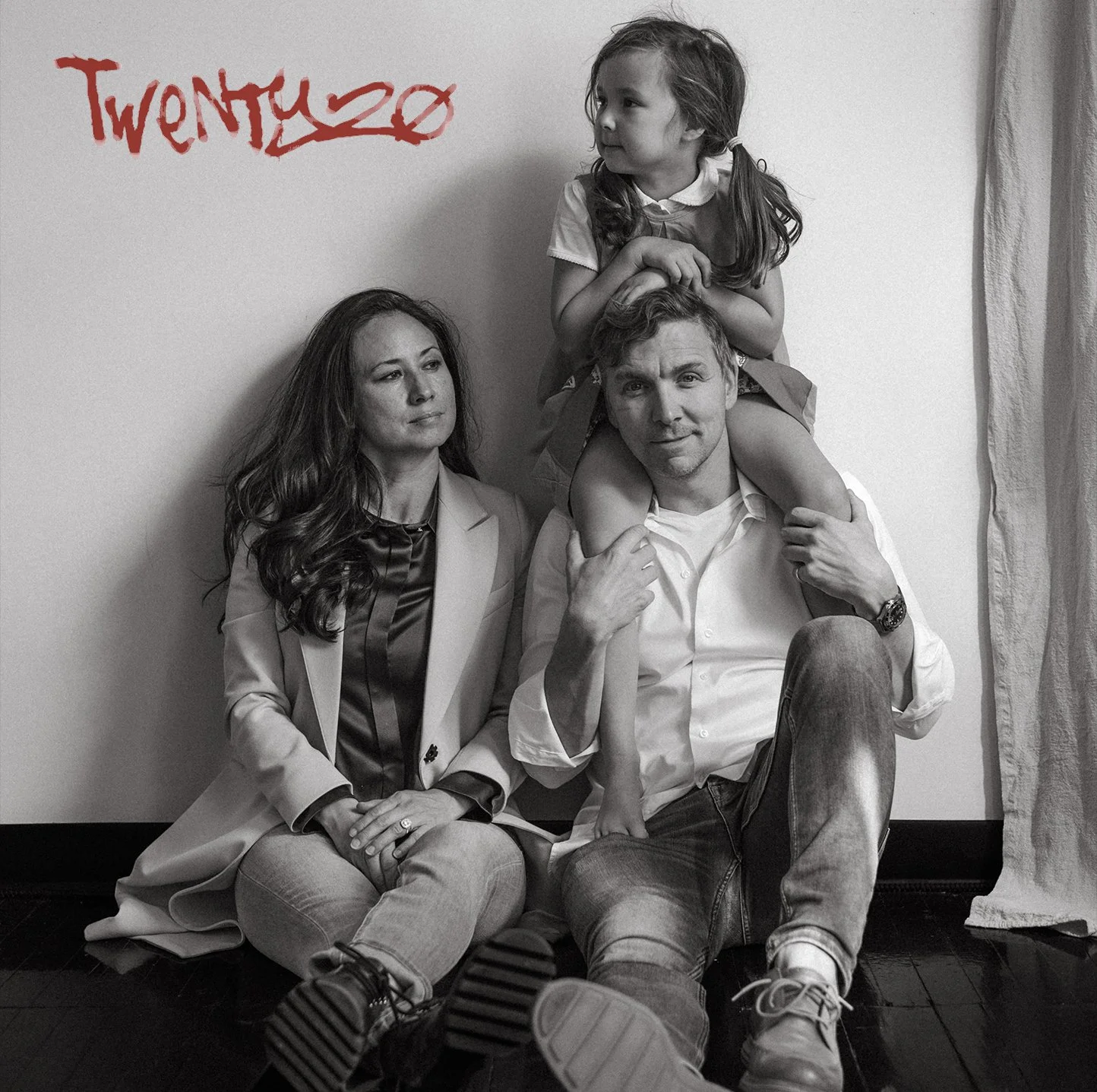 Black and white photo of a family sitting against a wall, with a young girl sitting on the father's shoulders. The family members are posing calmly, with the mother and father sitting on the floor and the girl sitting on the father's shoulders. The word "Twen Ty 20" is written in red on the wall.