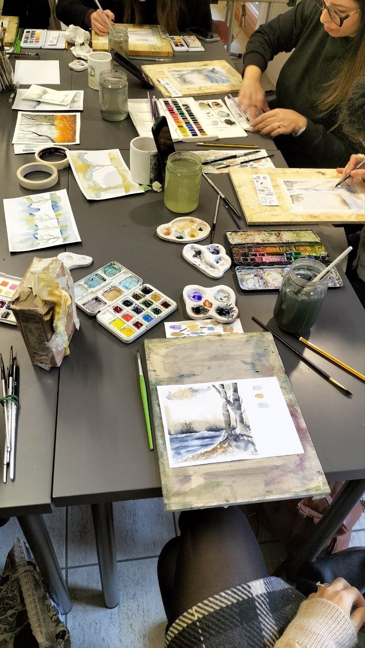 People sitting around a table painting watercolor landscapes with various painting supplies, including watercolors, brushes, jars of water, and paper.