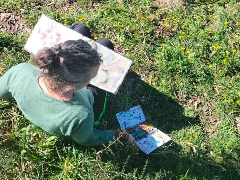 Woman sitting at a wooden table painting a colorful abstract artwork with a brush, surrounded by art supplies, with a lamp and art materials on a cluttered desk behind her.