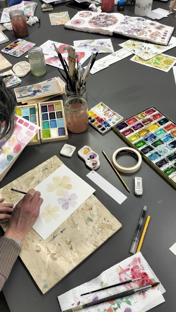 Waterscolors, paintbrushes, and floral watercolor paintings under progress on a worktable, surrounded by art supplies and sketches.