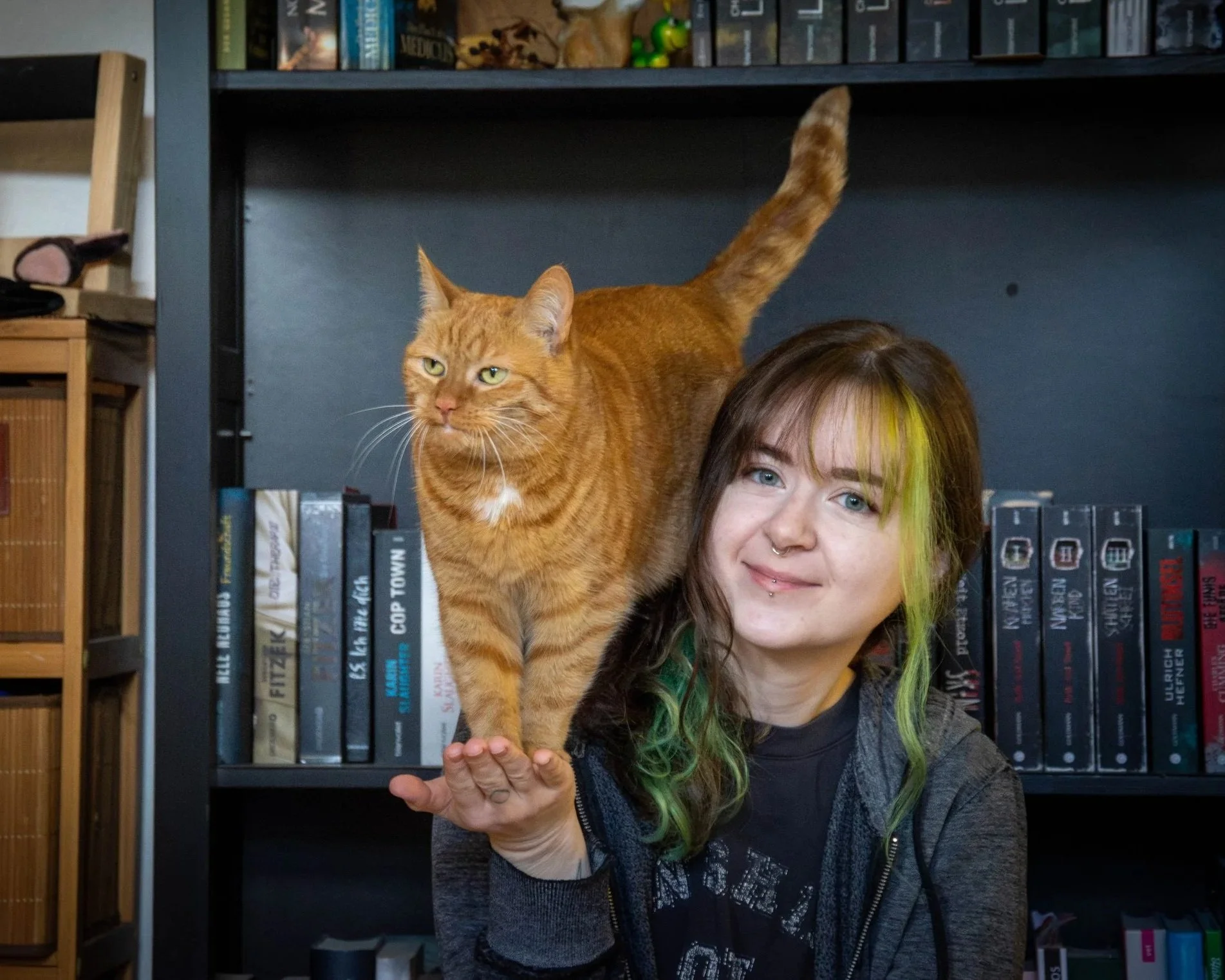 Junge Frau mit grünen Haarspitzen und Nasenpiercing hält einen orangefarbenen Kater vor einer Bücherwand.