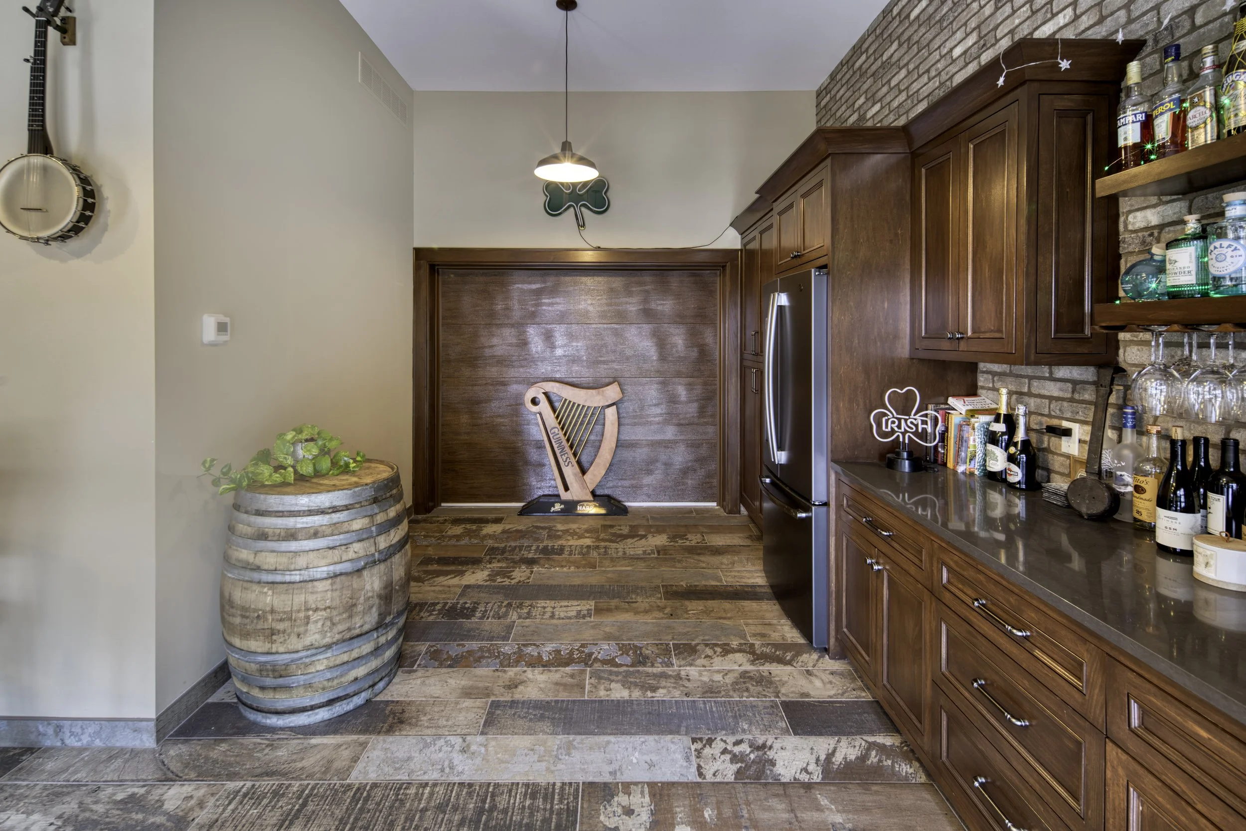 A kitchen area with wooden cabinets, a stainless steel refrigerator, liquor bottles on a shelf, and a barrel with a plant on top, with a wooden harp-shaped Guinness sign on the wall.