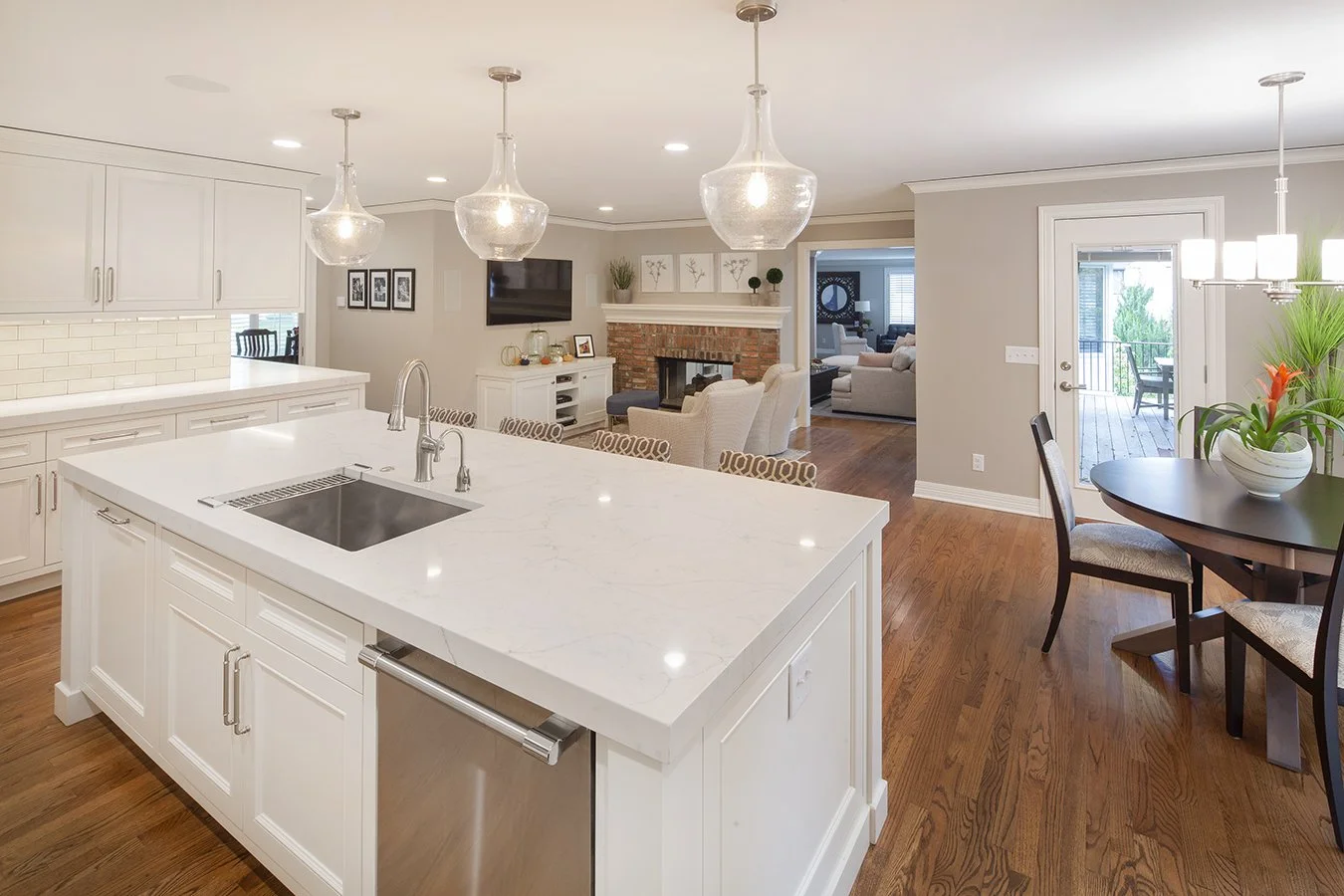 Open-concept kitchen and living room with white cabinets, marble island, hardwood flooring, brick fireplace, and dining area with flowers in a white pot.