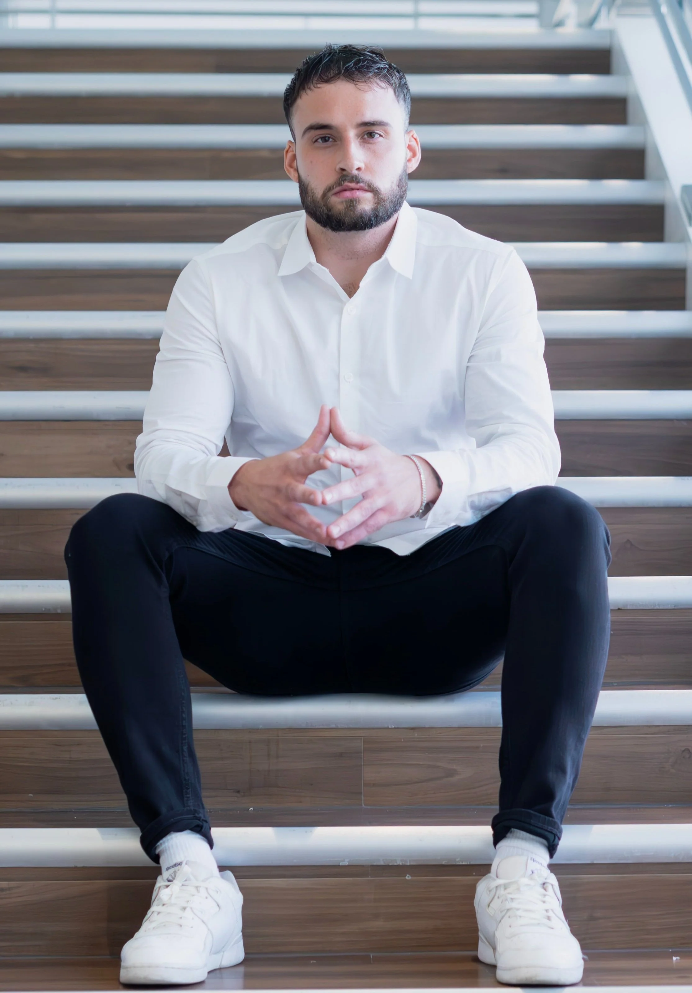 A man with dark hair and a beard sitting on wooden stairs, wearing a white shirt, black pants, and white sneakers, with his hands clasped in front of him.