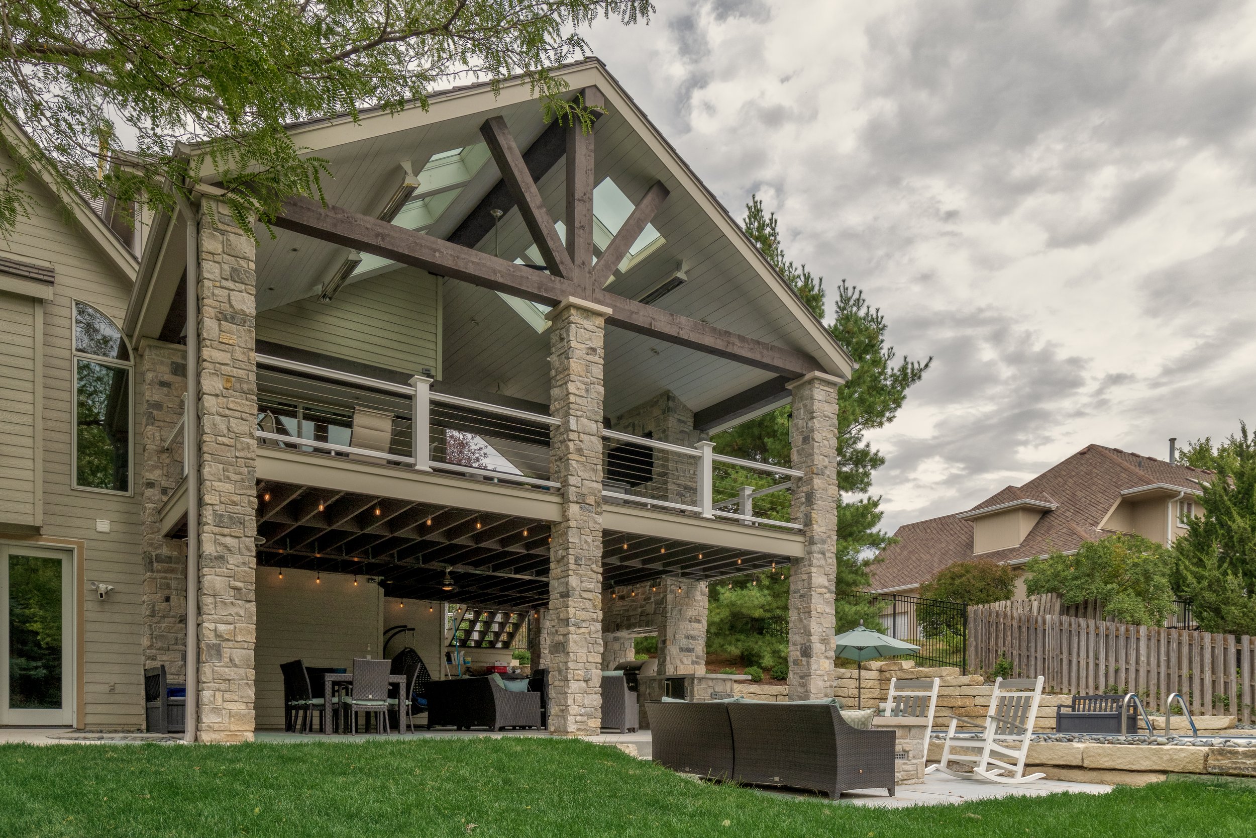 Backyard patio with two-story house with stone columns, seating area, umbrella, and a hot tub, surrounded by trees and a wooden fence.