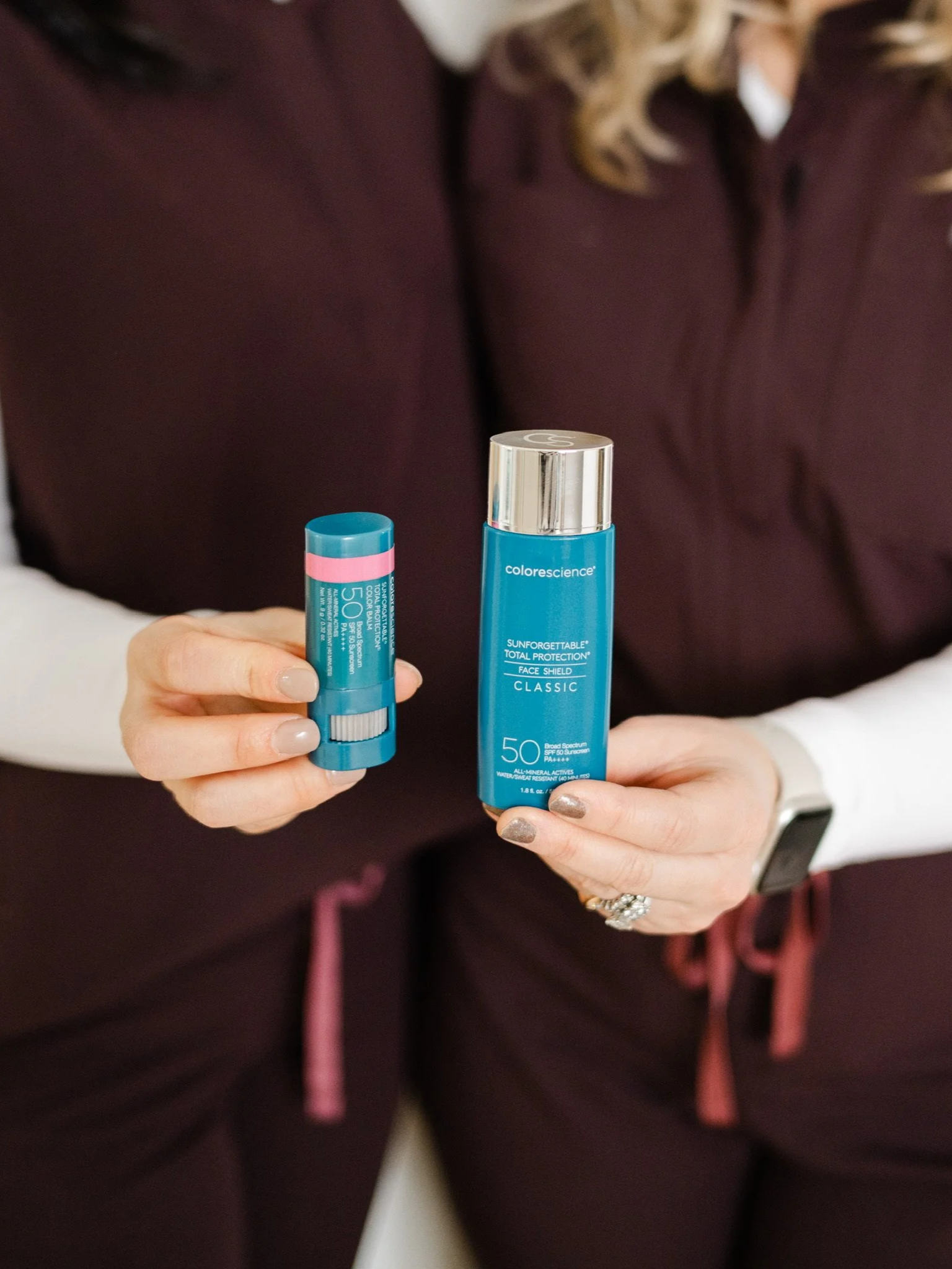 A woman holding a blue bottle of sunscreen and a smaller blue container of lip balm, both with SPF 50, in front of her torso.