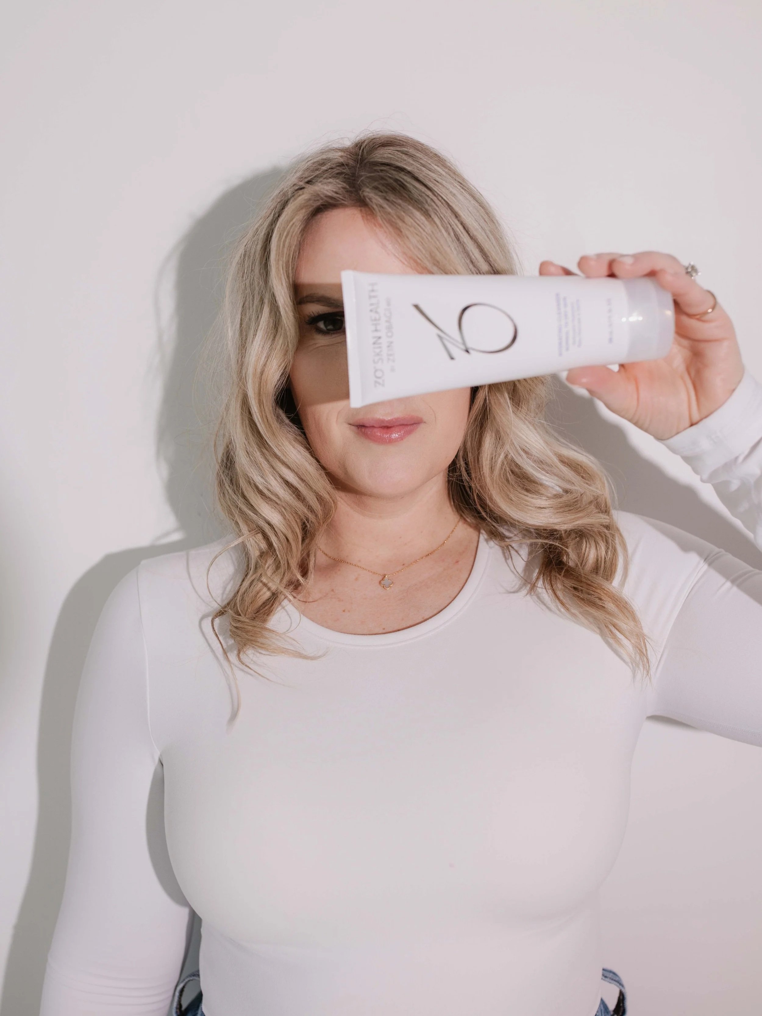A woman with blonde wavy hair, wearing a white long-sleeve shirt, holding a product in front of her face, covering one eye, against a white background.