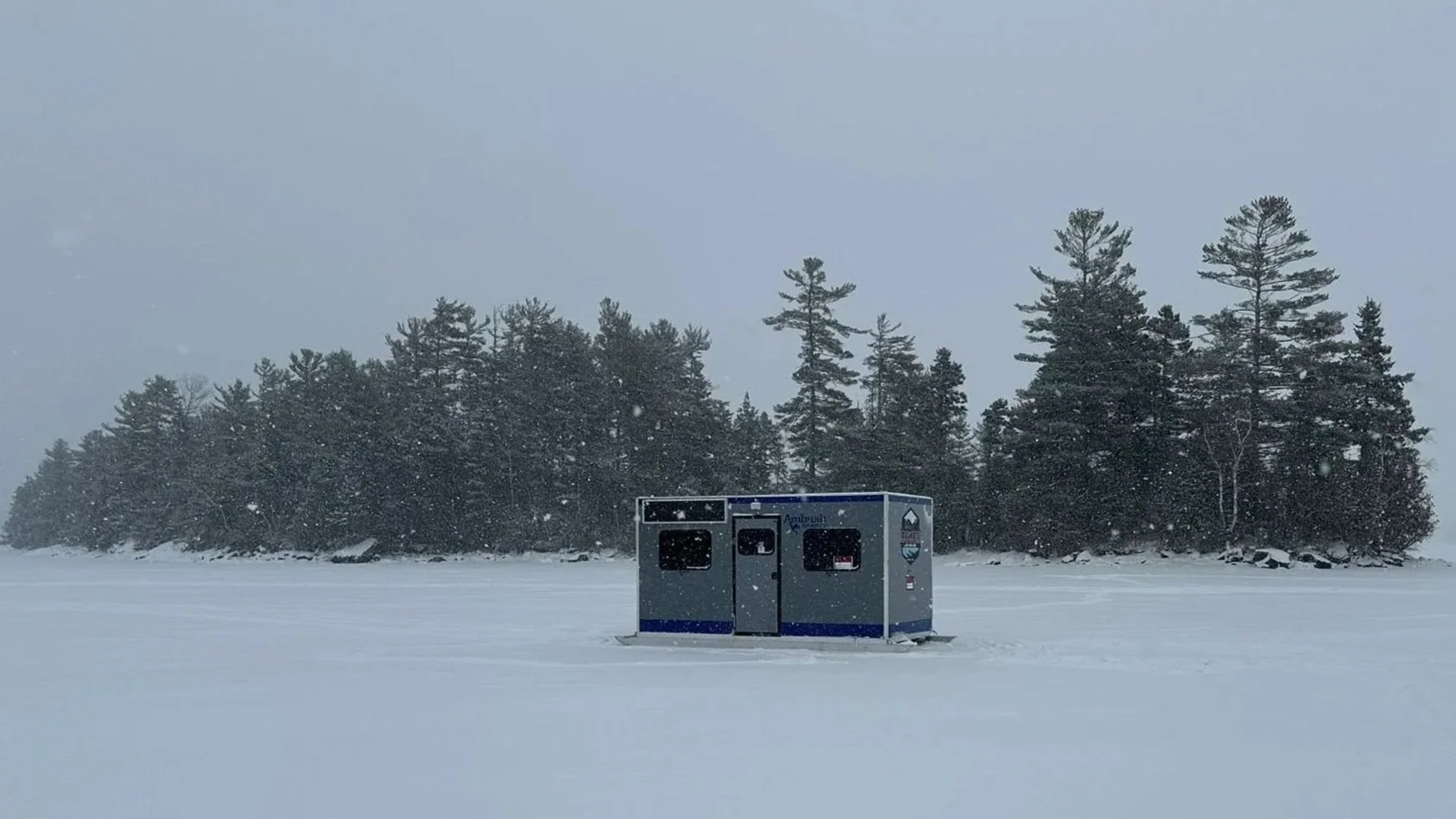A portable, silver-colored, rectangular camping trailer with two windows, a black vent, and branding that reads "Ambush SHANTY" on a grassy area outdoors.