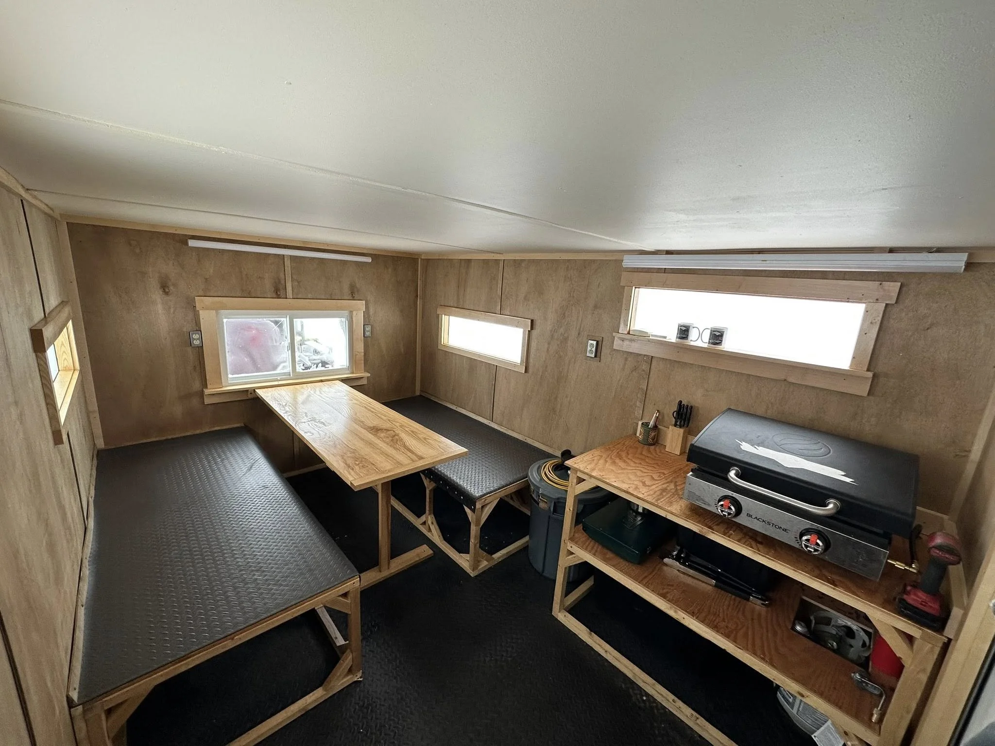 Interior of a small, finished room with three small windows, white walls, a plywood workbench in the center, and additional plywood benches along the sides.