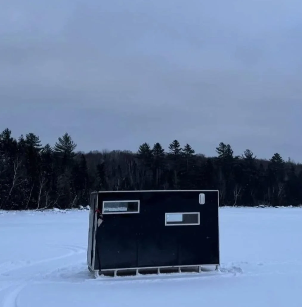 A small outdoor kitchen setup with a Blackstone griddle, a portable stove, and various utensils hanging on a wooden panel. There is a window and storage containers below.