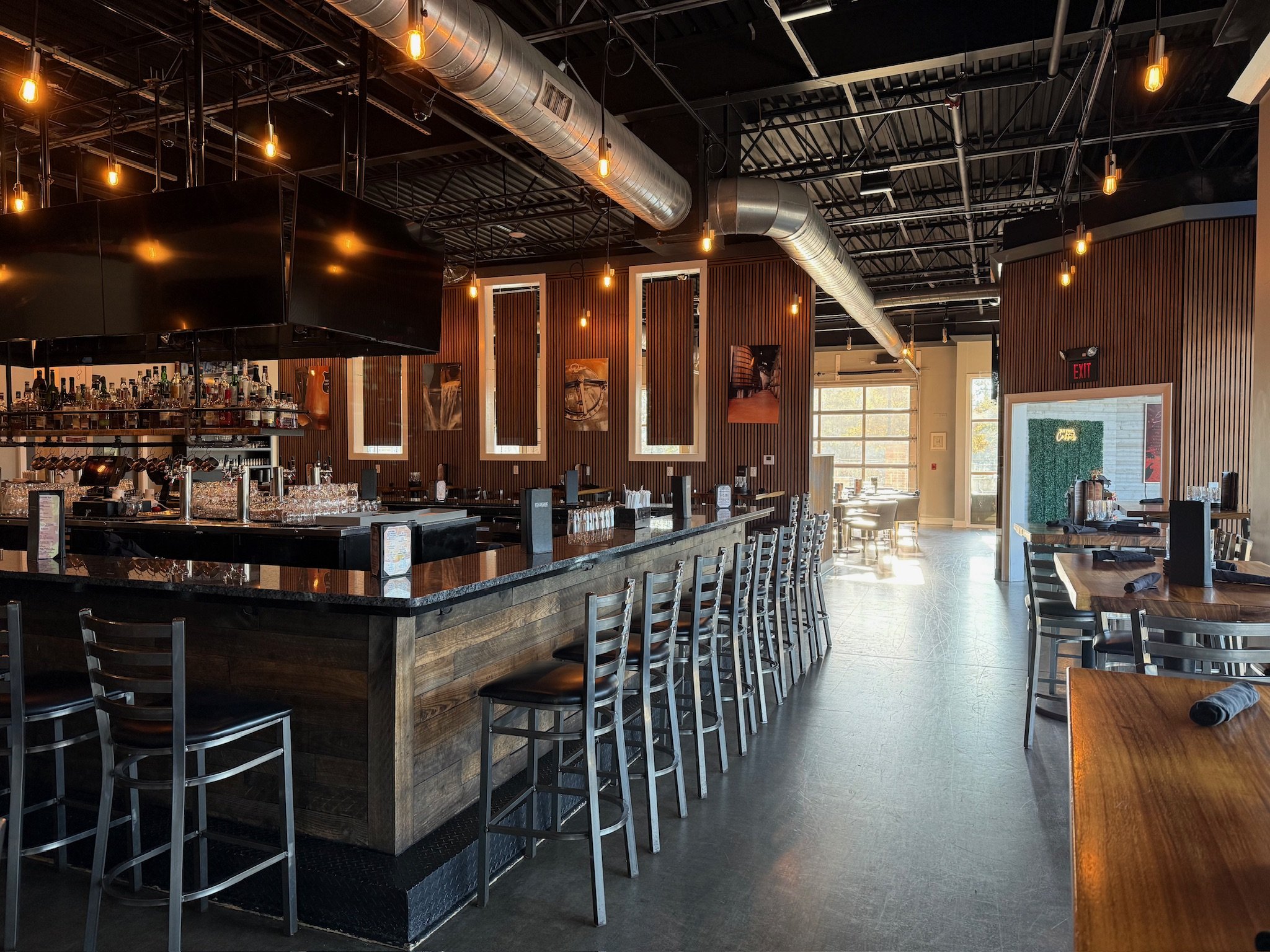 Interior of a modern restaurant or bar with a black ceiling, exposed ducts, warm lighting, wooden wall paneling, and a bar area with bottles and glasses.