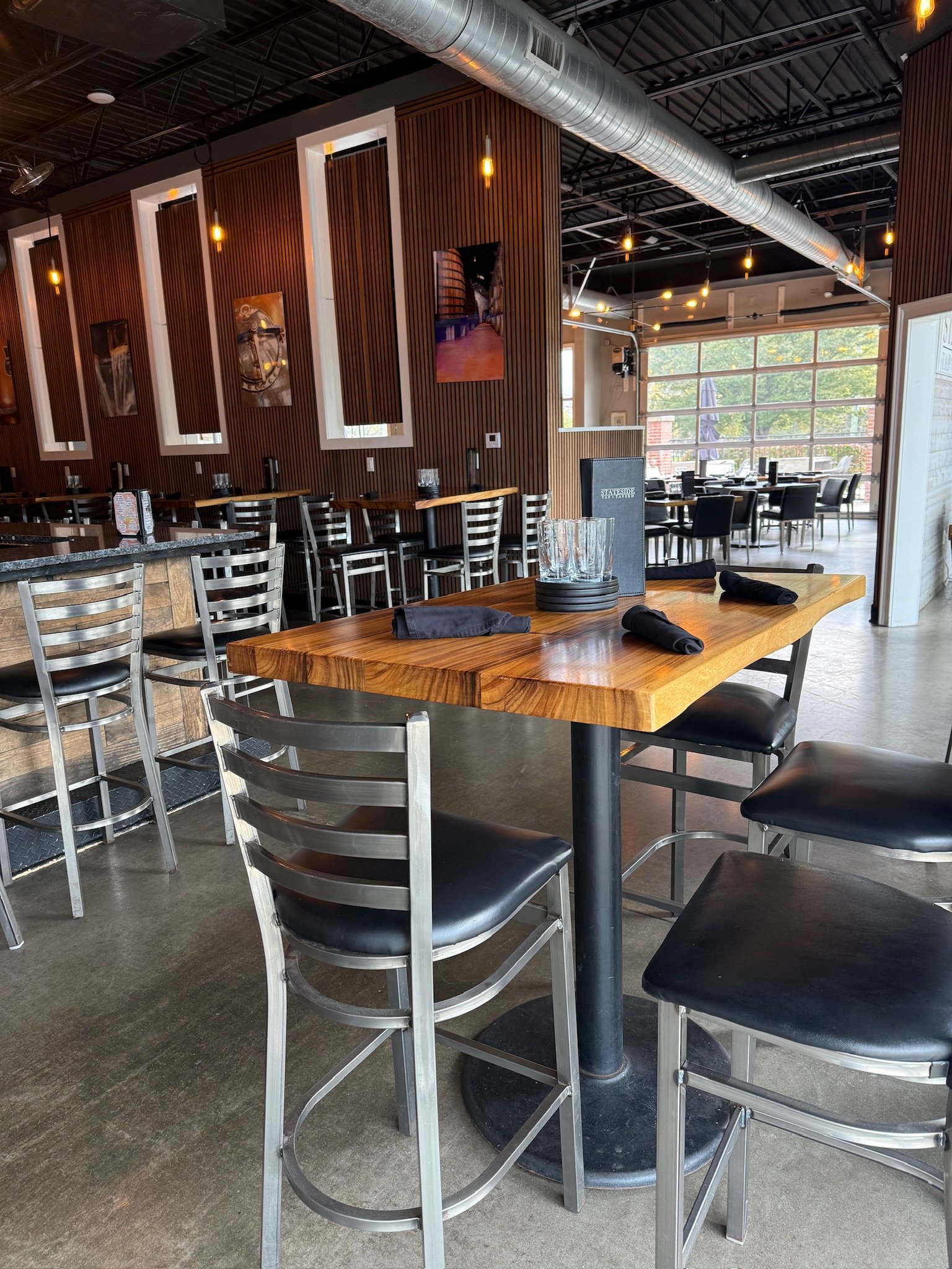 Interior of a modern restaurant or cafe with wooden wall panels, black chairs, a mix of high and low tables, large windows, and exposed industrial-style ceiling with ductwork and hanging lights.