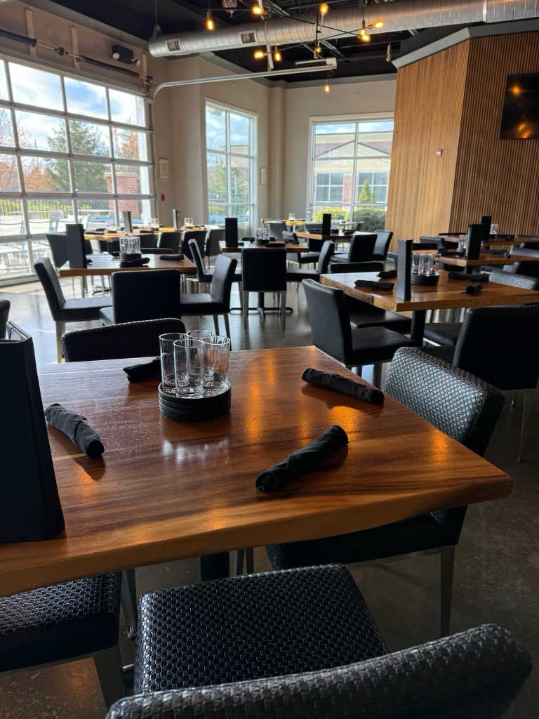 Interior of a modern restaurant or cafe with wooden tables, black chairs, glasses, and cloth napkins, illuminated by hanging Edison bulbs with large windows showing an outdoor patio.