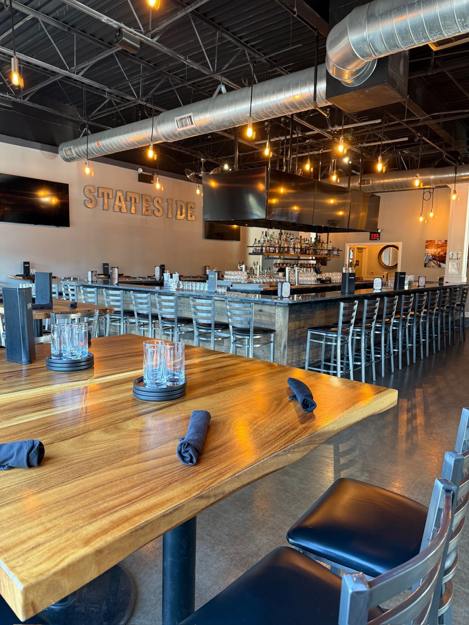 Interior of a modern restaurant with wooden tables and chairs, a bar area with bottles of alcohol, industrial-style lighting and ventilation, and the word 'STATESIDE' on the wall.