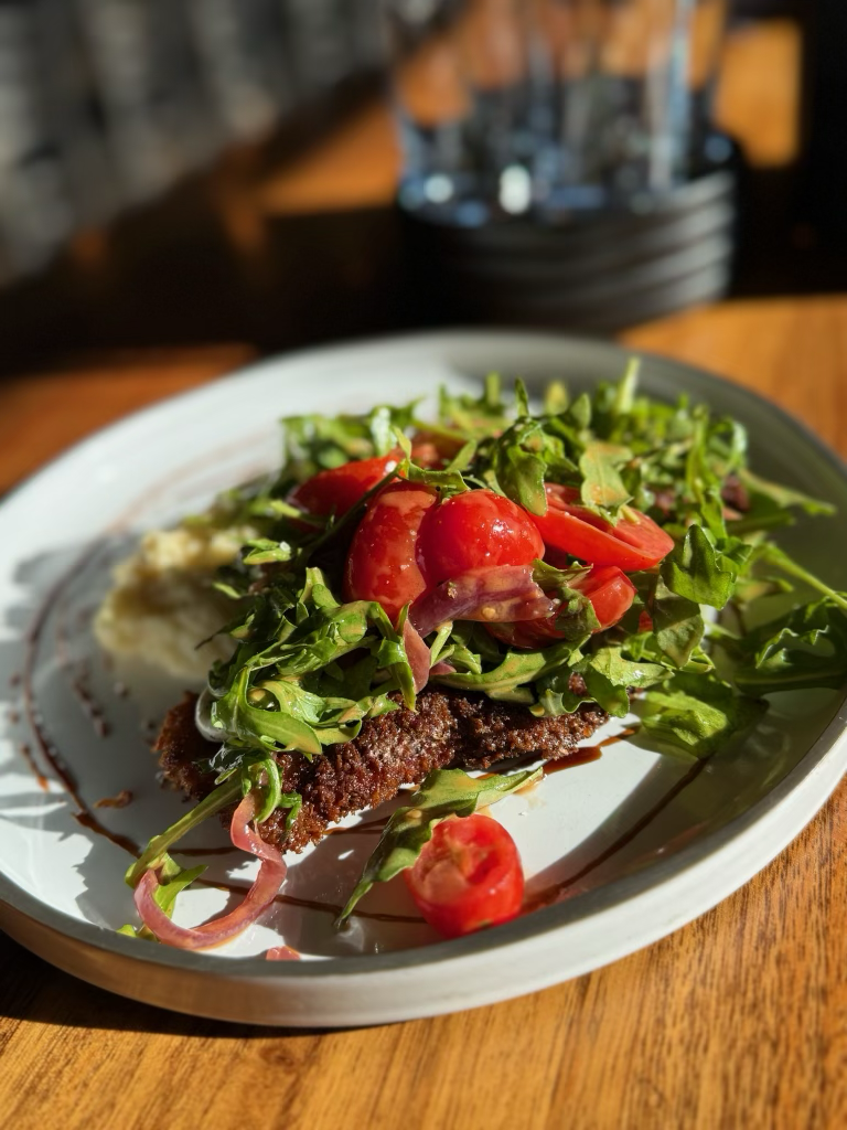 Plate of fried meat topped with fresh salad greens, cherry tomatoes, and sliced vegetables, garnished with a drizzle of sauce, on a rustic wooden table.