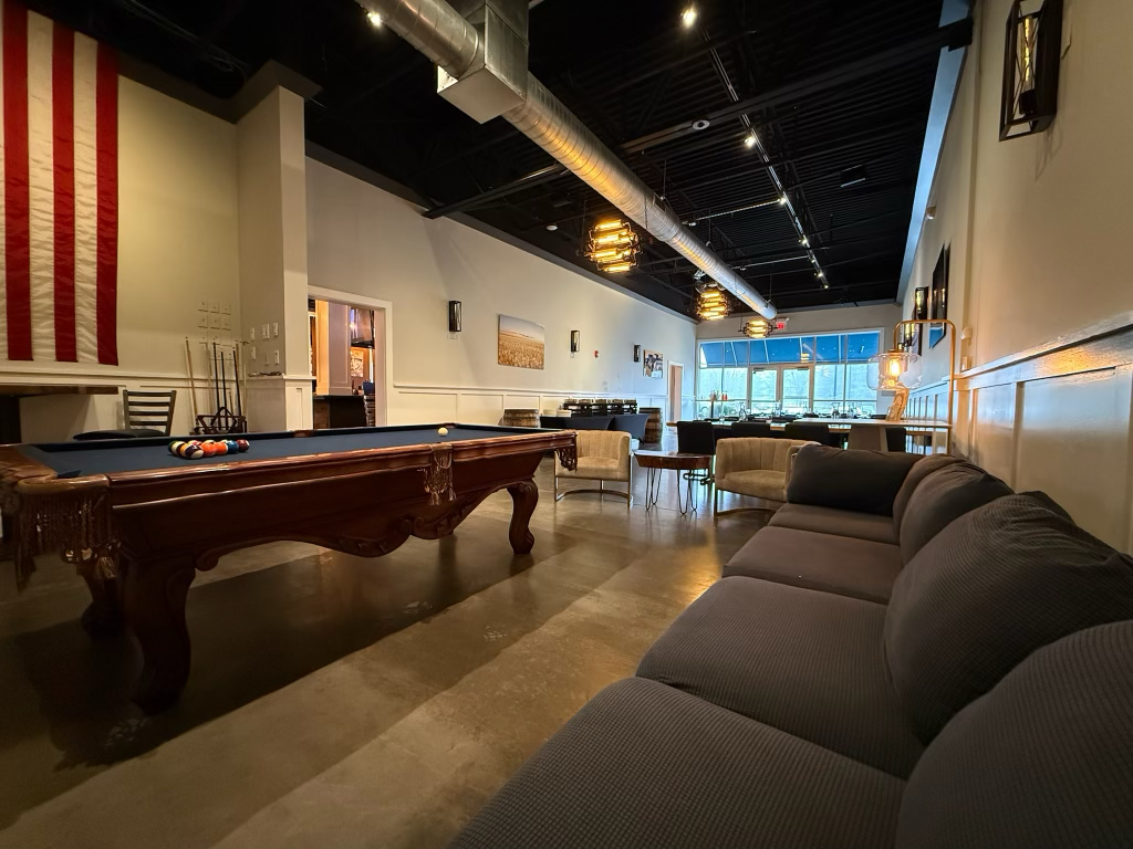 Interior of a lounge with a pool table, a beige sofa, armchairs, and large windows, decorated with modern light fixtures and wall art.