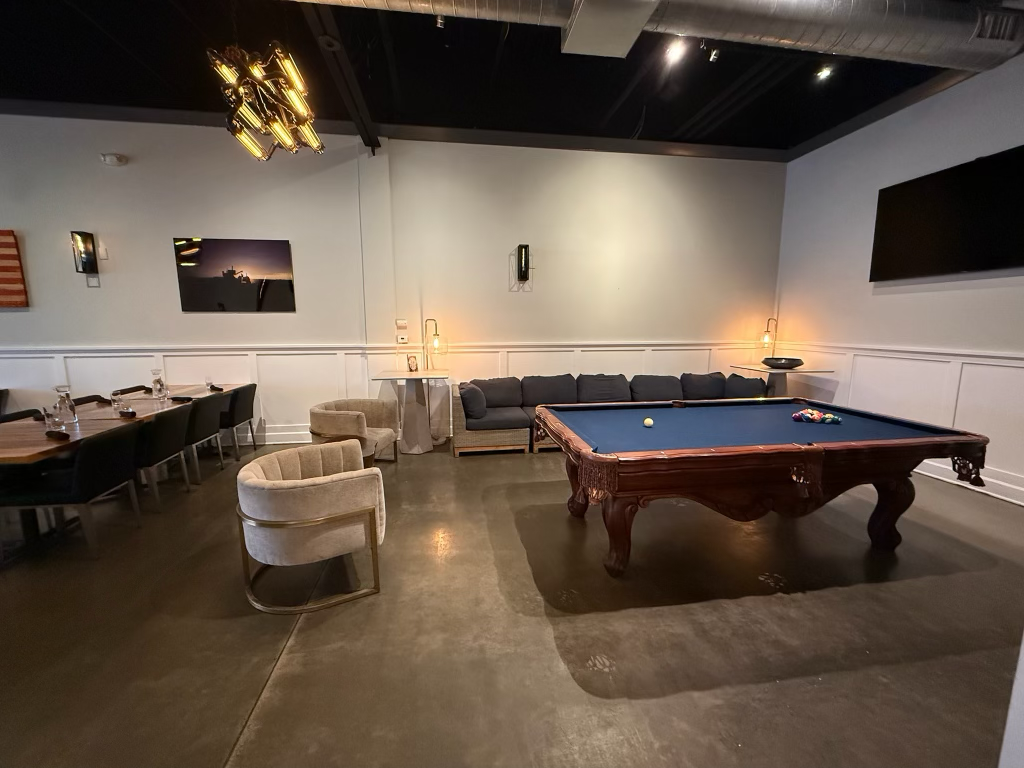 A game room with a pool table, a black sectional sofa, several beige chairs, a long dining table with black chairs, wall art, and lighting fixtures.