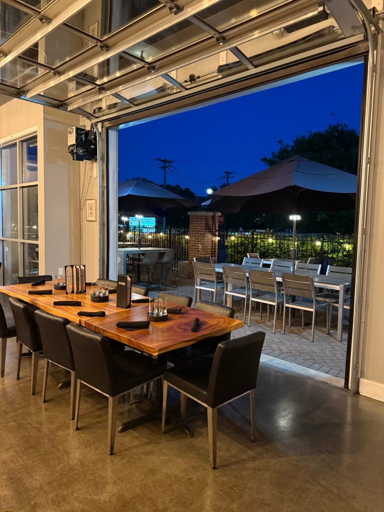 Indoor restaurant dining area with a wooden table set for six, black chairs, and a view of outdoor patio with tables, chairs, and umbrellas at night.