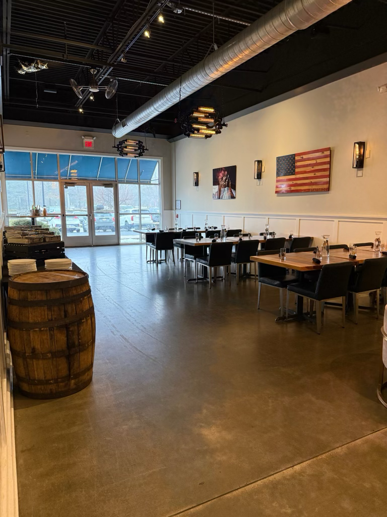 Empty restaurant with black tables and chairs, American flag artwork on the wall, large window at the front, and industrial ceiling fixtures.