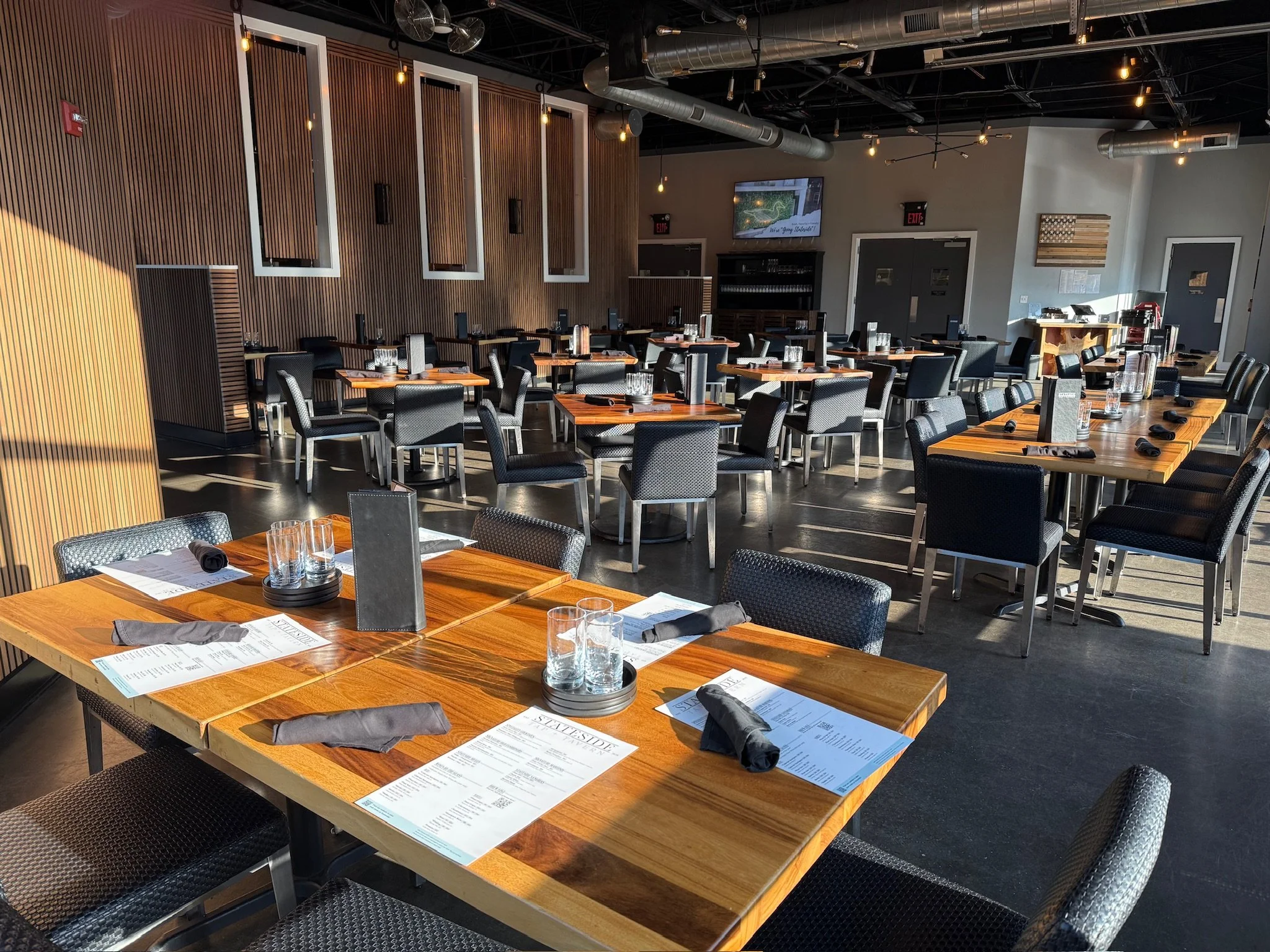 Interior of a modern restaurant with wooden tables set with glasses, menus, and black napkins, black chairs, a wooden accent wall, and a TV screen on the wall.