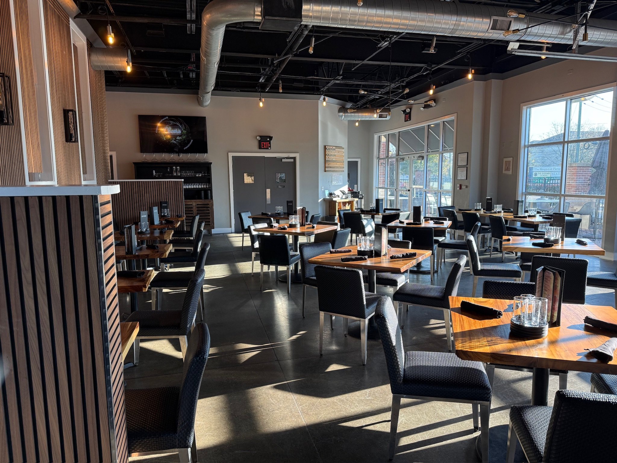 Interior of a modern restaurant with wooden tables, black chairs, large windows, exposed ductwork, and warm lighting.