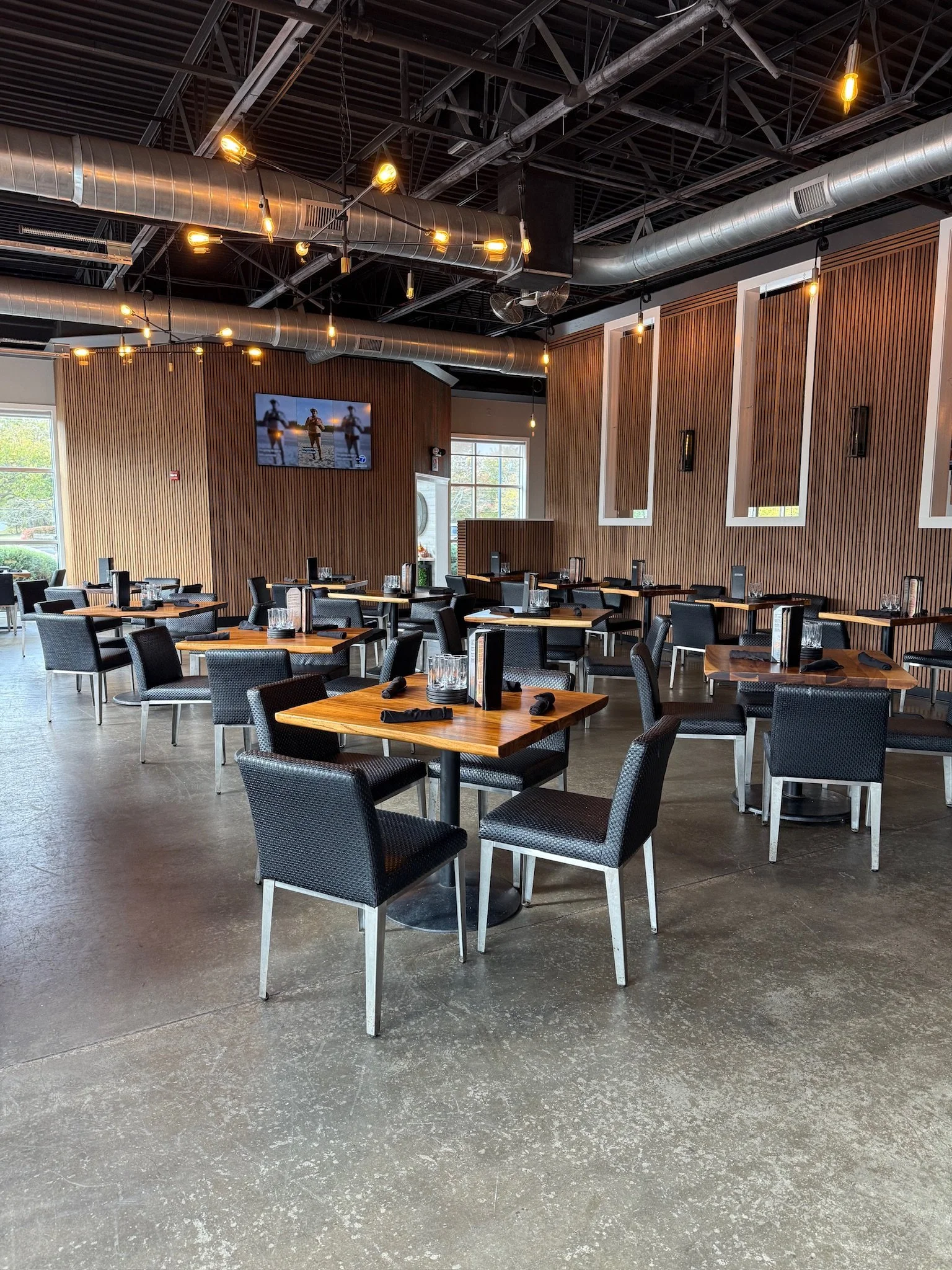 Interior of a modern restaurant with wooden panel walls, black chairs, and tables set with glasses, napkins, and menus. There are large windows, a TV on the wall, and industrial-style exposed ductwork and lighting.