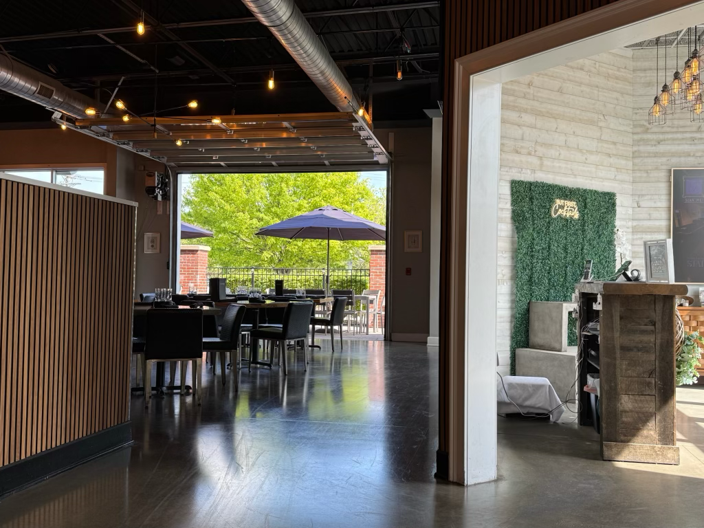 Inside view of a modern restaurant with an open patio, featuring tables, chairs, and umbrellas, with greenery outside.
