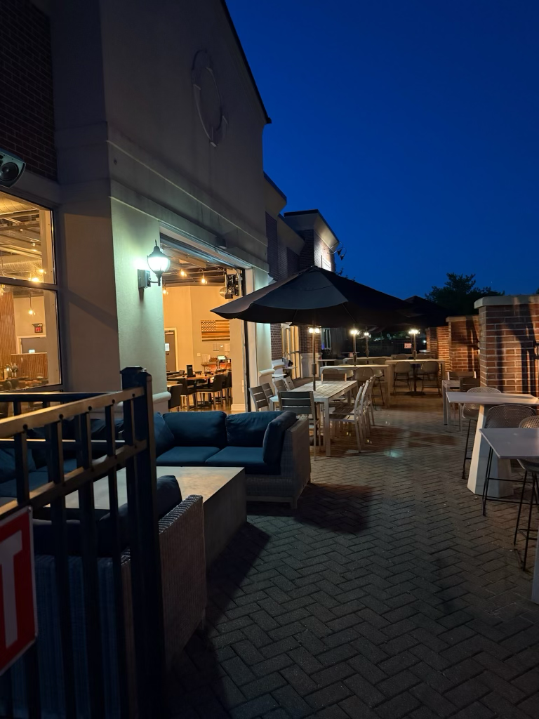 Outdoor patio area of a restaurant or cafe at night with tables, chairs, umbrellas, and a building with large windows and warm interior lighting.