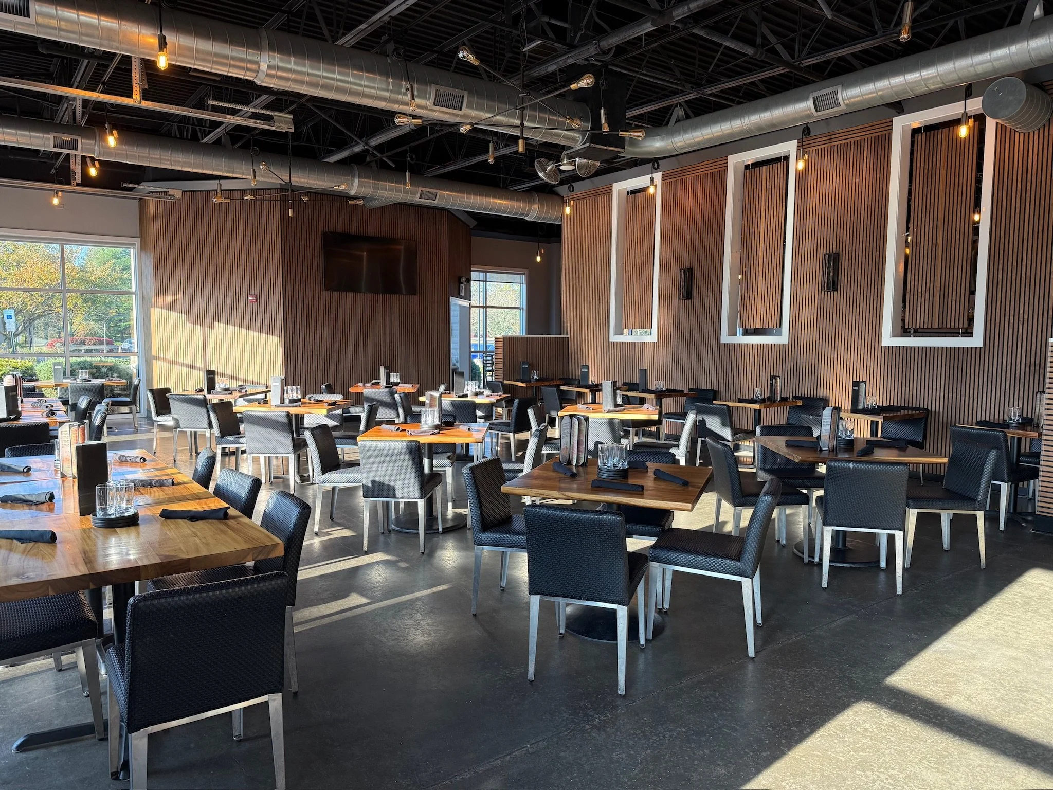 Interior of a modern restaurant or café with wooden wall paneling, black chairs, tables set with glasses and napkins, large windows letting in natural light, and an industrial-style ceiling with exposed ductwork and hanging Edison bulbs.