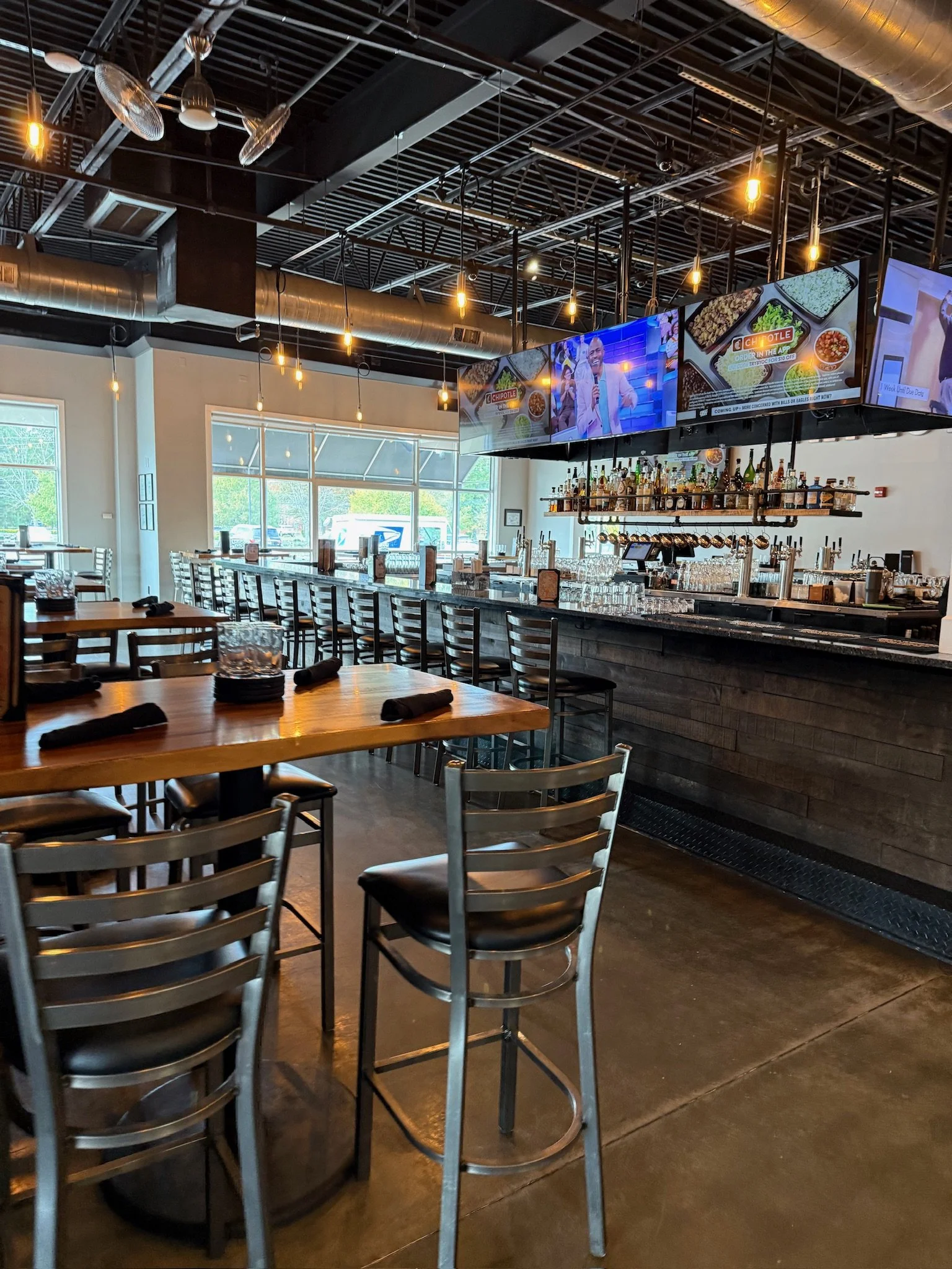 Interior of a modern restaurant or bar with a marble countertop, rows of bar stools, wooden tables with black cloth napkins, hanging Edison light bulbs, and digital screens showing a man and food advertisements.
