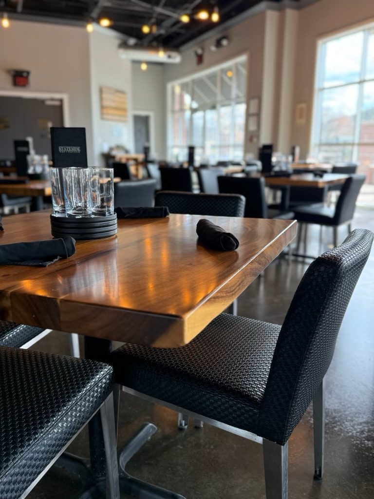 Empty restaurant table with black chairs, glasses, and black napkins, near large windows with sunlight in a modern dining area.