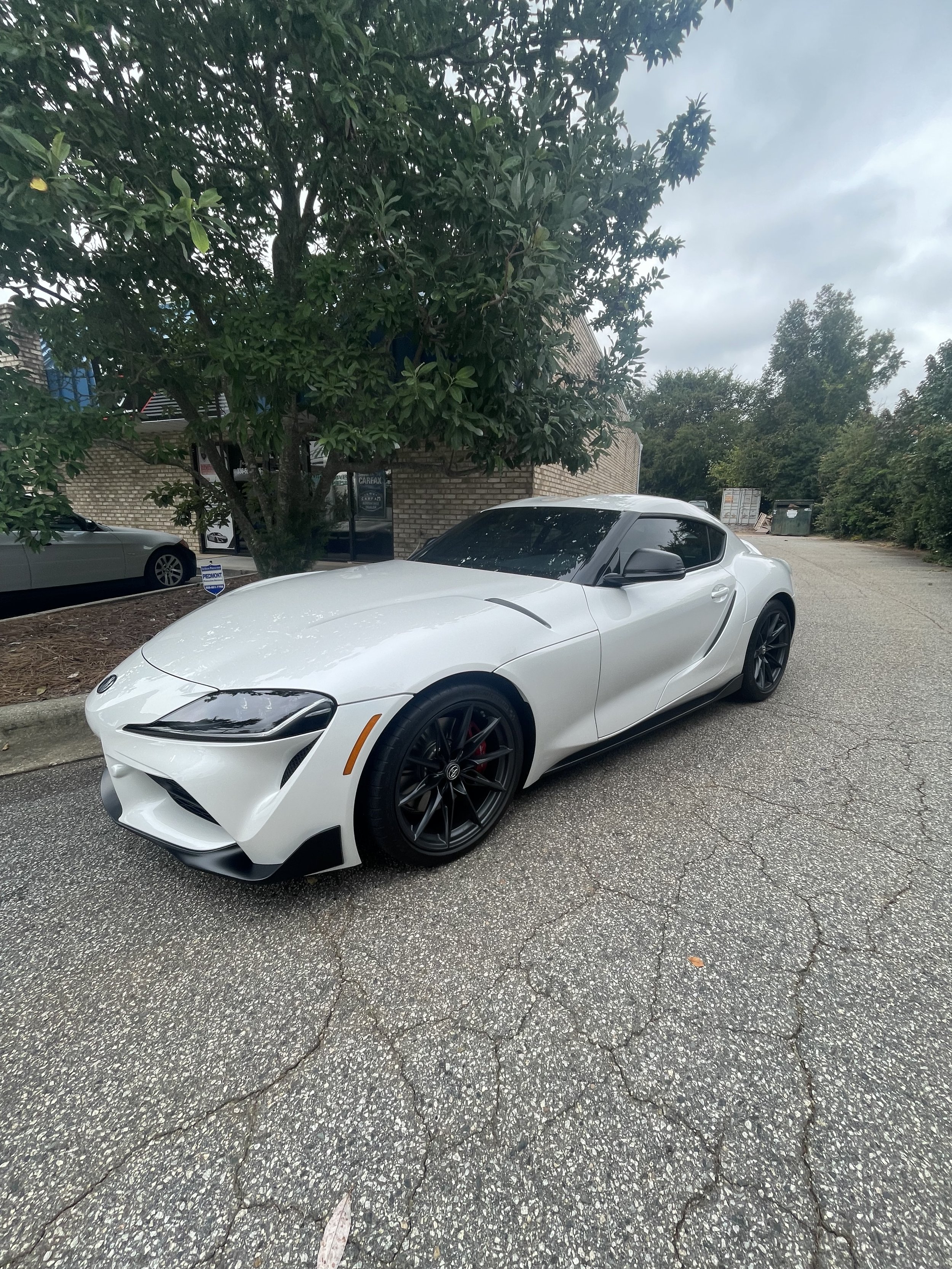White sports car parked with window tint on asphalt, surrounded by greenery and buildings.