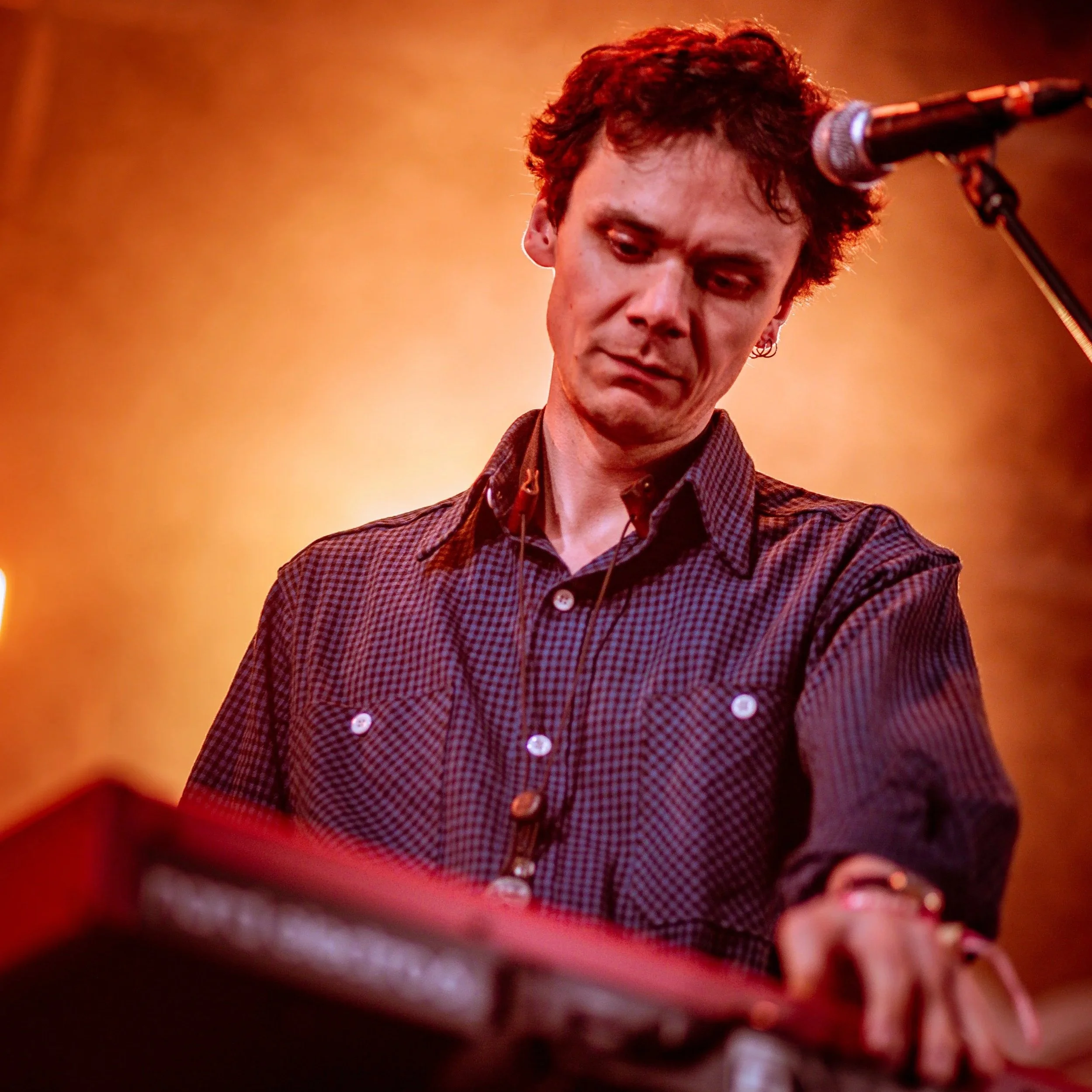 A man with dark, curly hair wearing a checkered shirt, focused on playing a keyboard or electronic instrument in a performance setting with warm stage lighting.