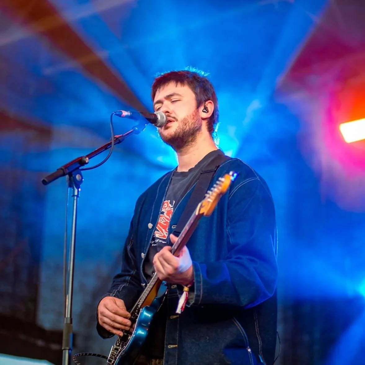 Male musician singing and playing electric guitar on stage with colorful stage lights.